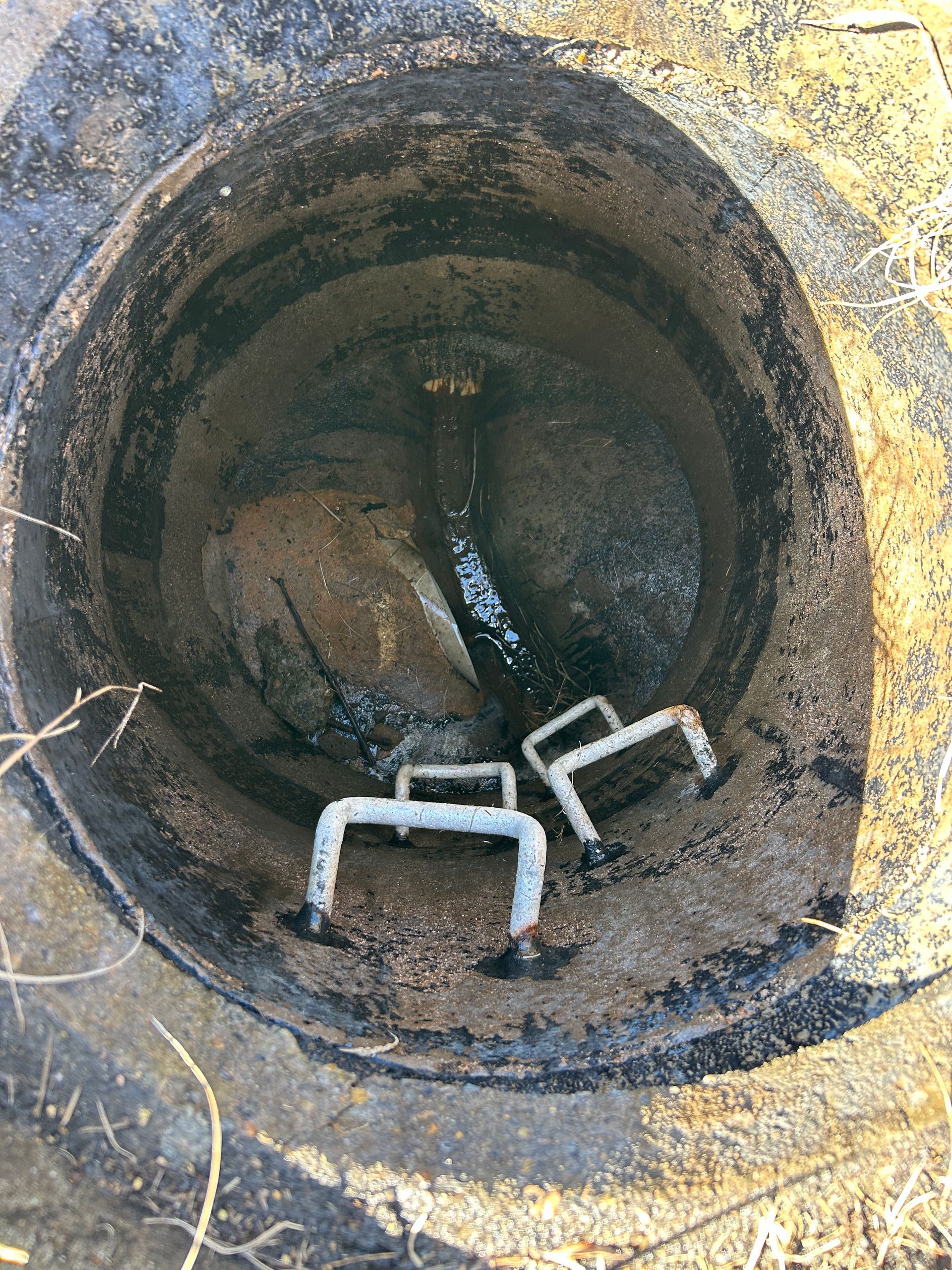 An open, dark circular manhole revealing a concrete interior with metal rungs for climbing and a narrow drainage pipe.