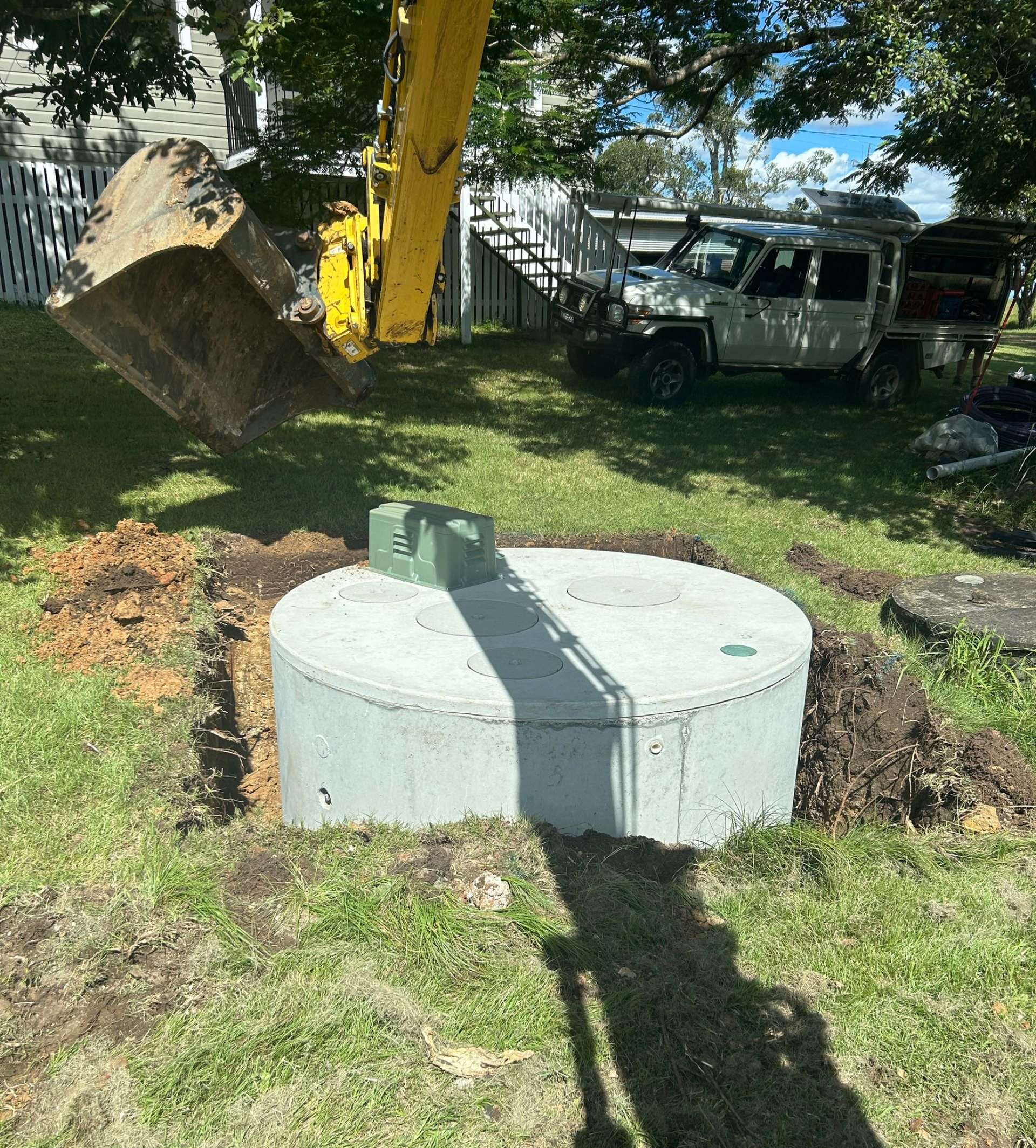 An excavator bucket hovers over a newly installed circular concrete septic tank in a residential yard.