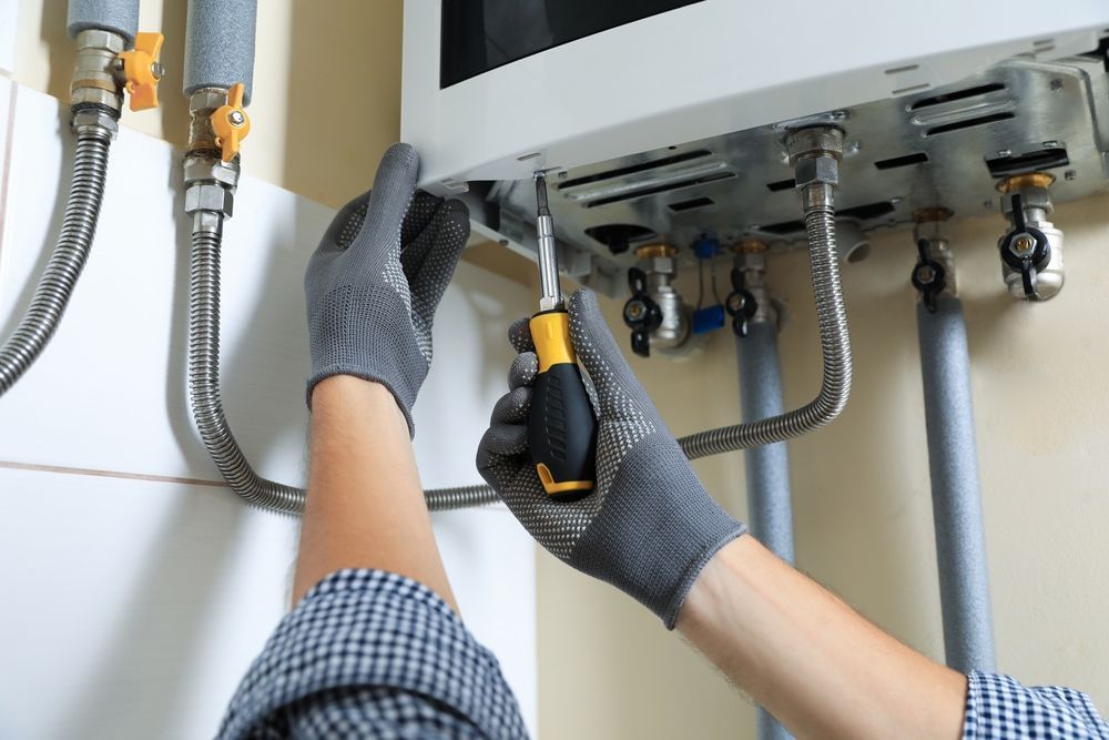 A technician wearing grey gloves uses a screwdriver to service a white wall-mounted boiler.