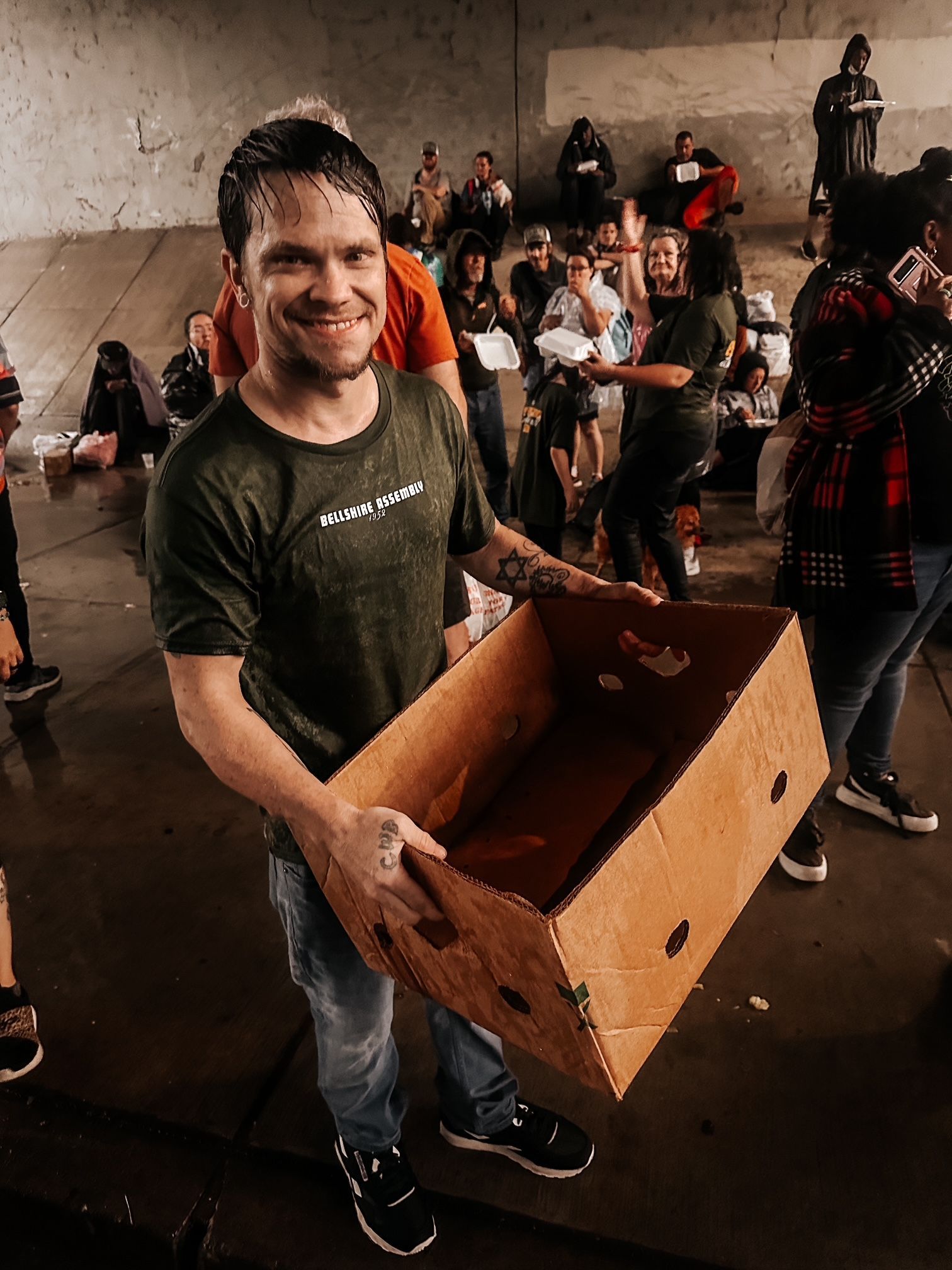 A person smiling while holding a cardboard box in a crowded, dimly lit indoor space where others are receiving supplies.