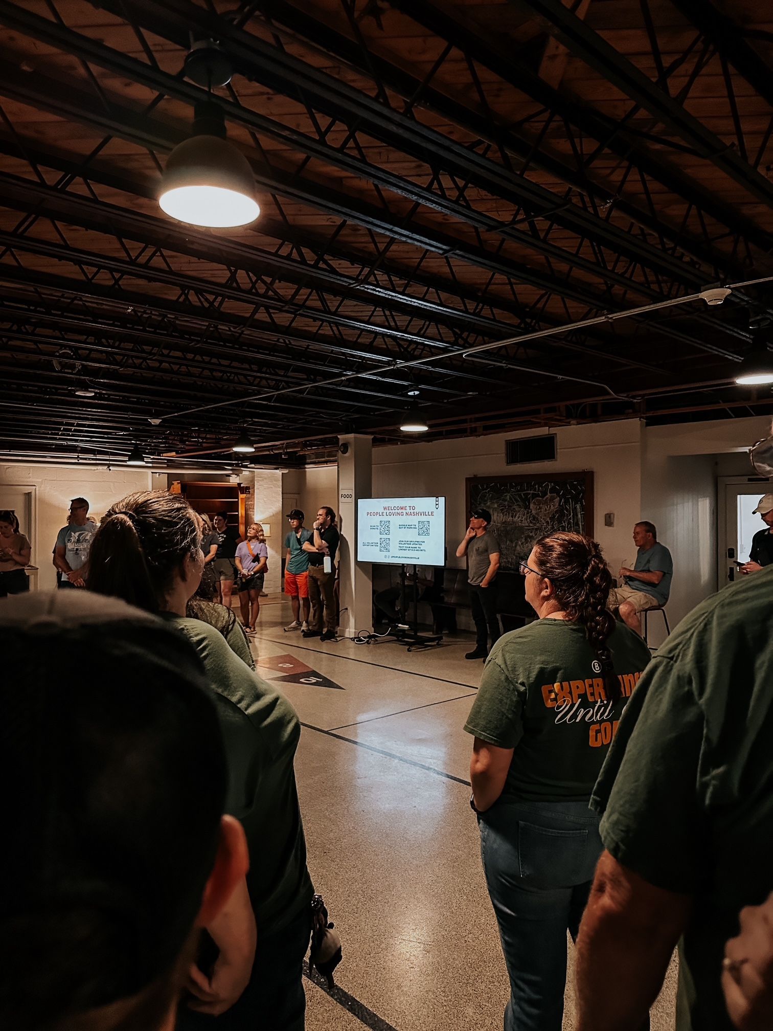 A group of people standing in a dimly lit, rustic room with exposed rafters, listening to a presentation on a screen.