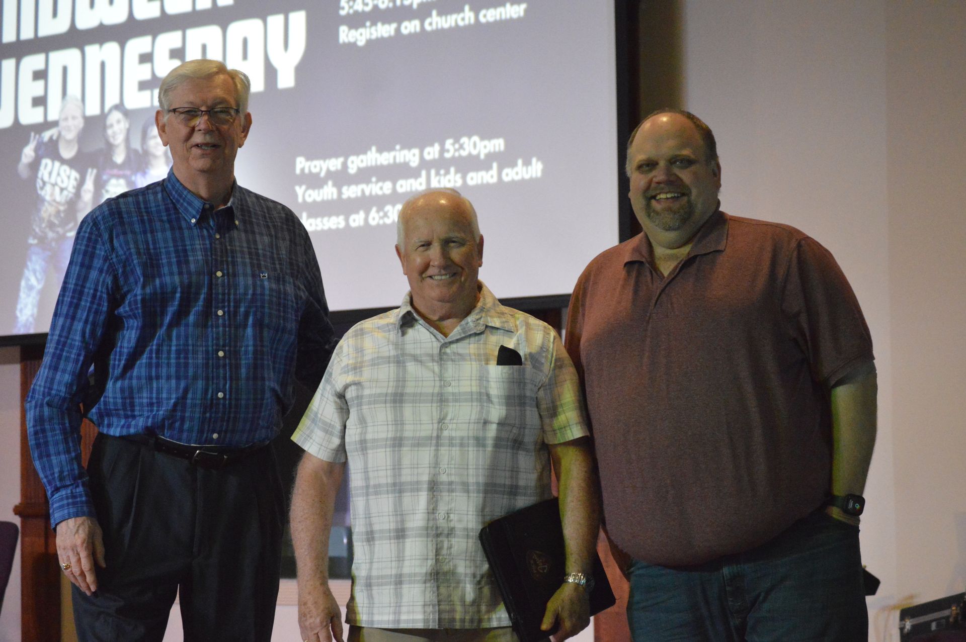 Three men stand smiling in front of a projection screen about church activities.
