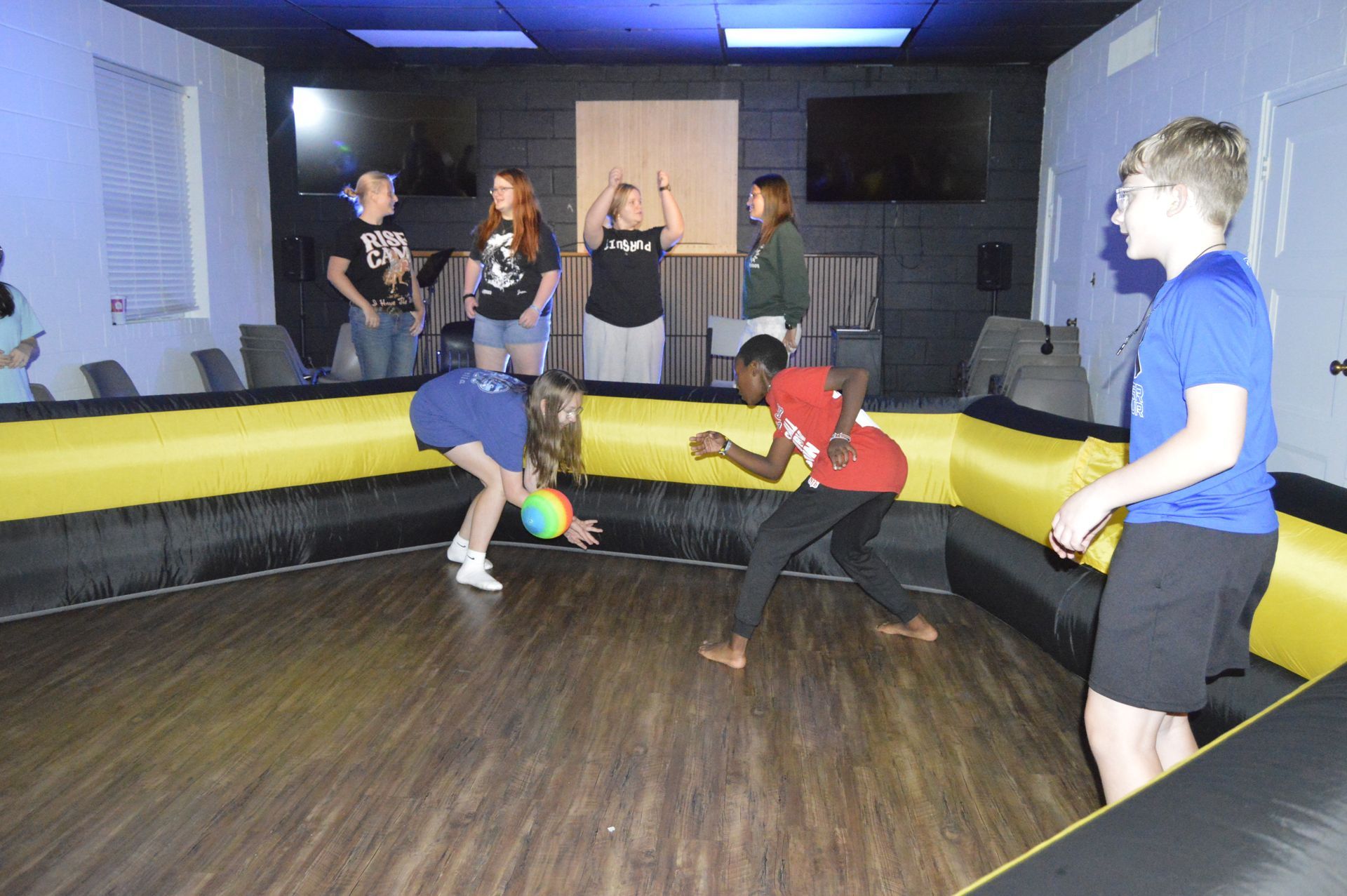 People playing a game in an inflatable-walled arena inside a room with wood-look flooring and overhead lighting.