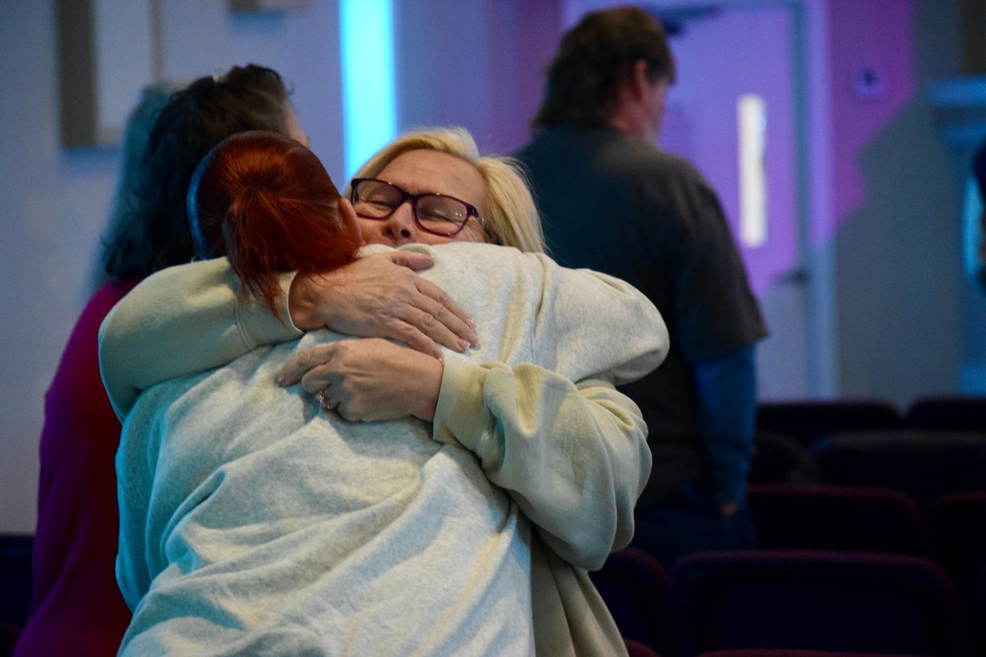 Two people embrace in a dimly lit room with rows of chairs in the background.