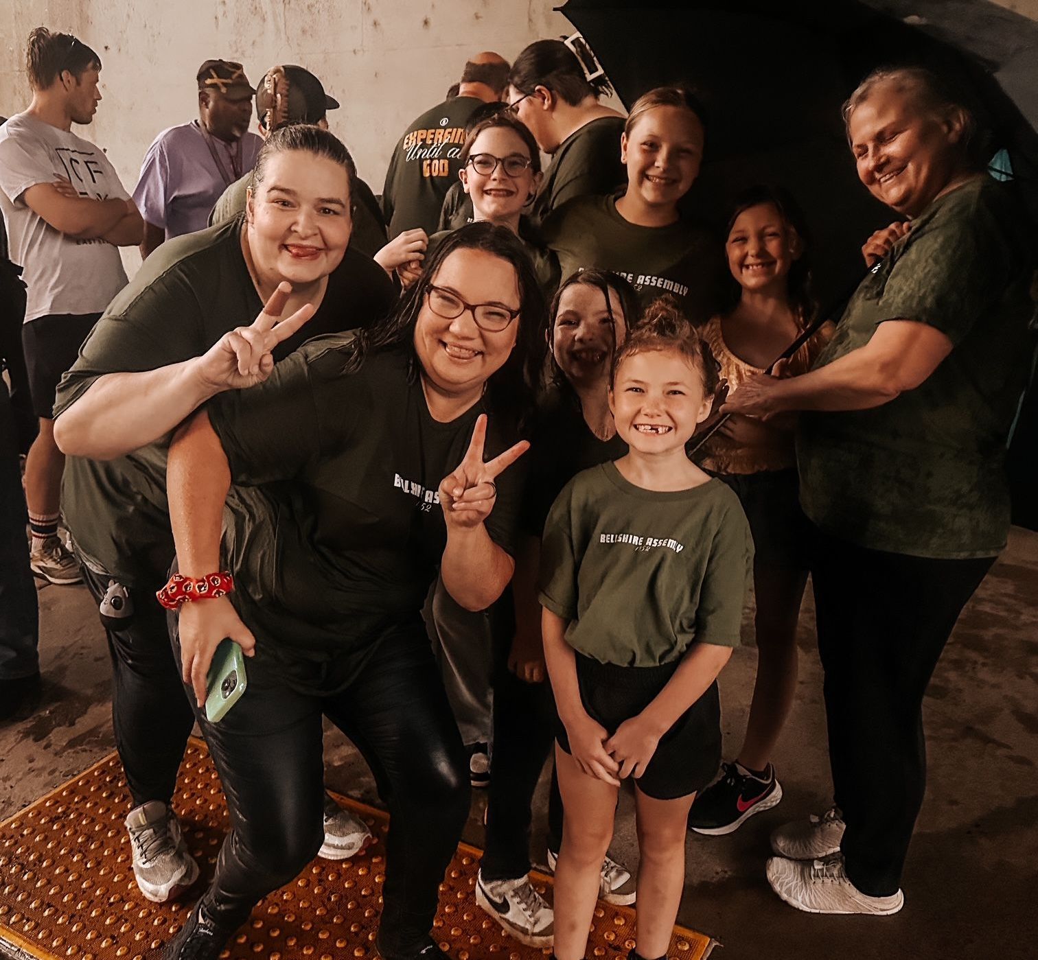 A group of people wearing matching olive green shirts gather for a photo indoors, many smiling and making peace signs.