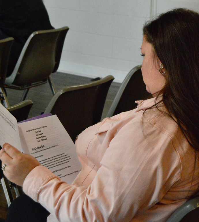 A person with long dark hair, wearing a light pink shirt, sits in a chair reading a paper program in an indoor space.