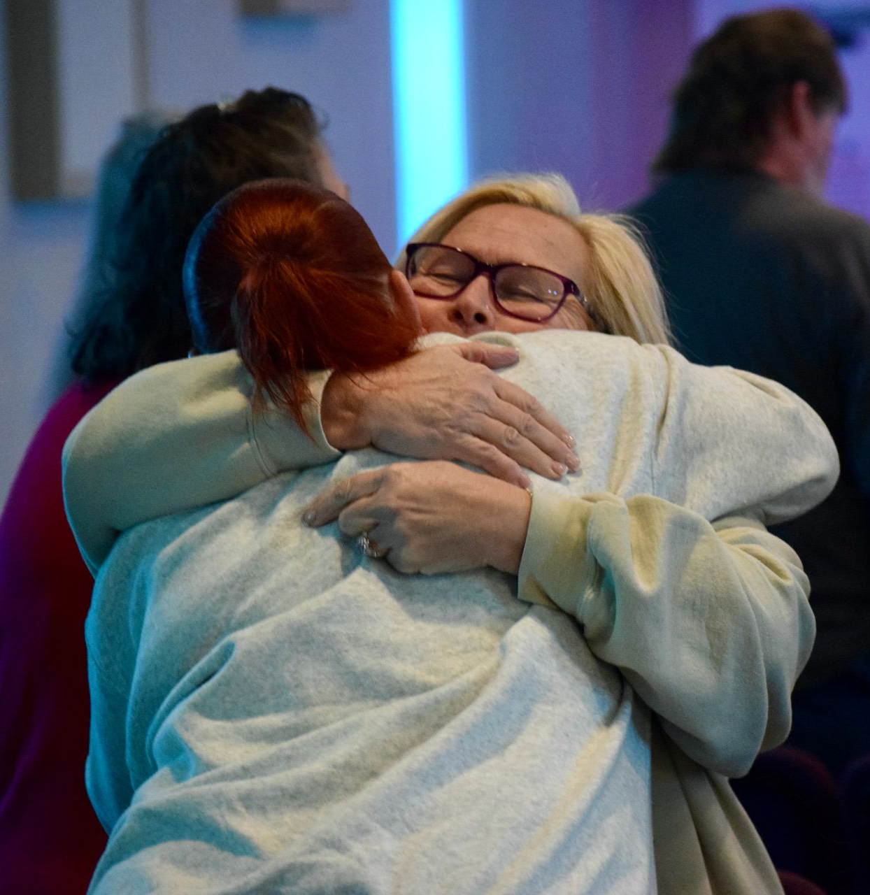 Two people embrace, one with blonde hair wearing glasses and a light-colored sweater, the other with auburn hair.