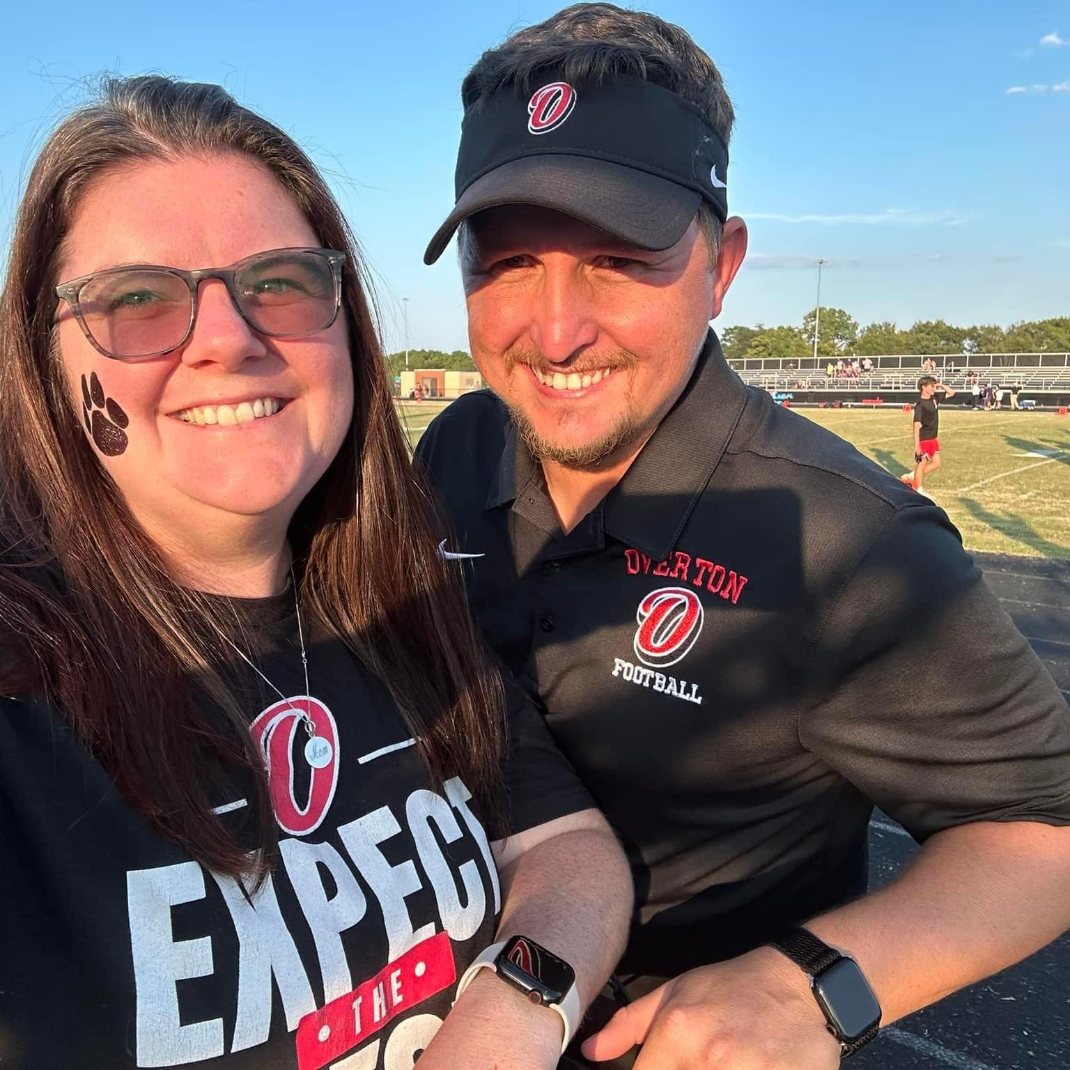 Two people in matching black sports team apparel smile for a selfie on a sunny outdoor athletic field.