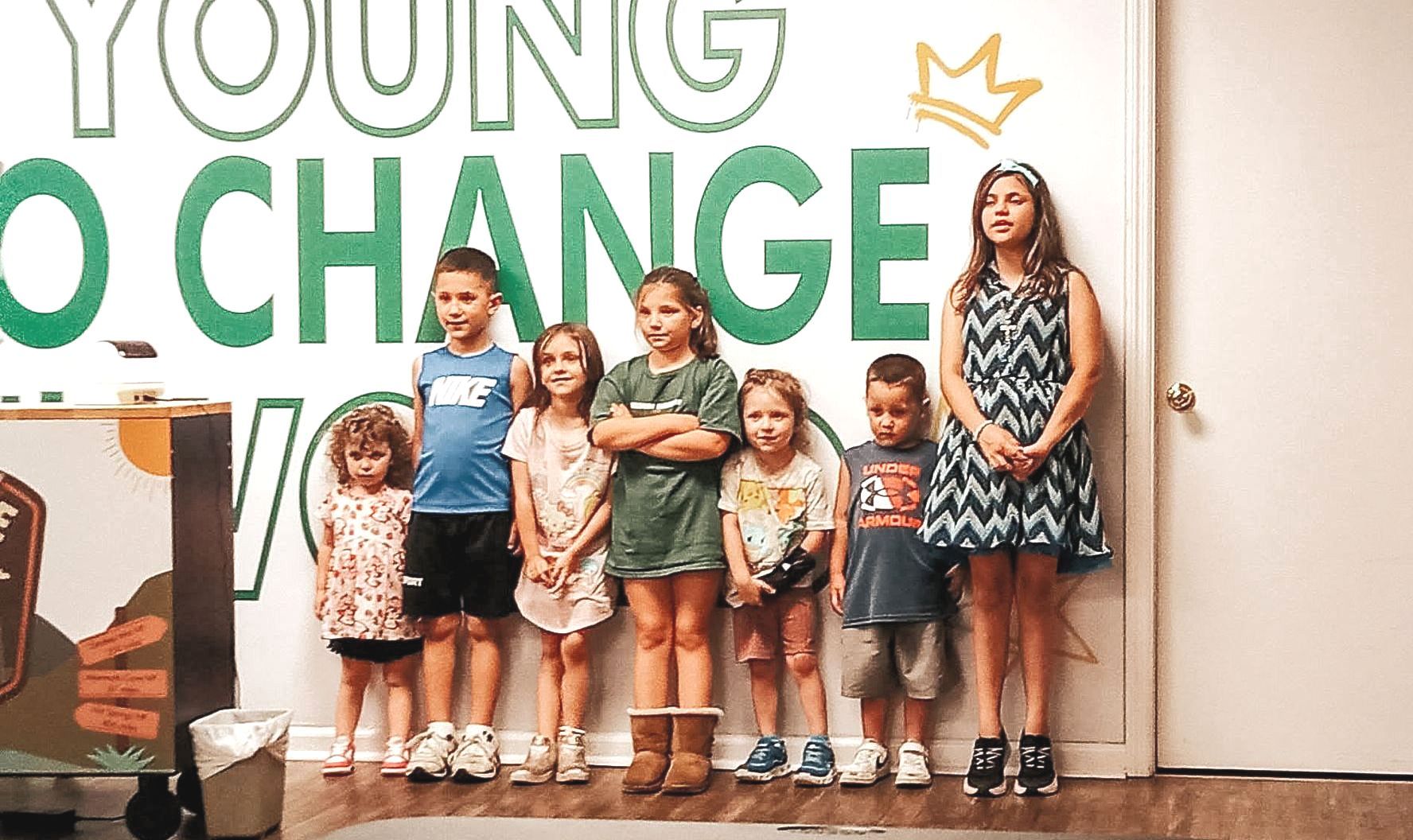 Seven children standing in a row against a wall with the words 