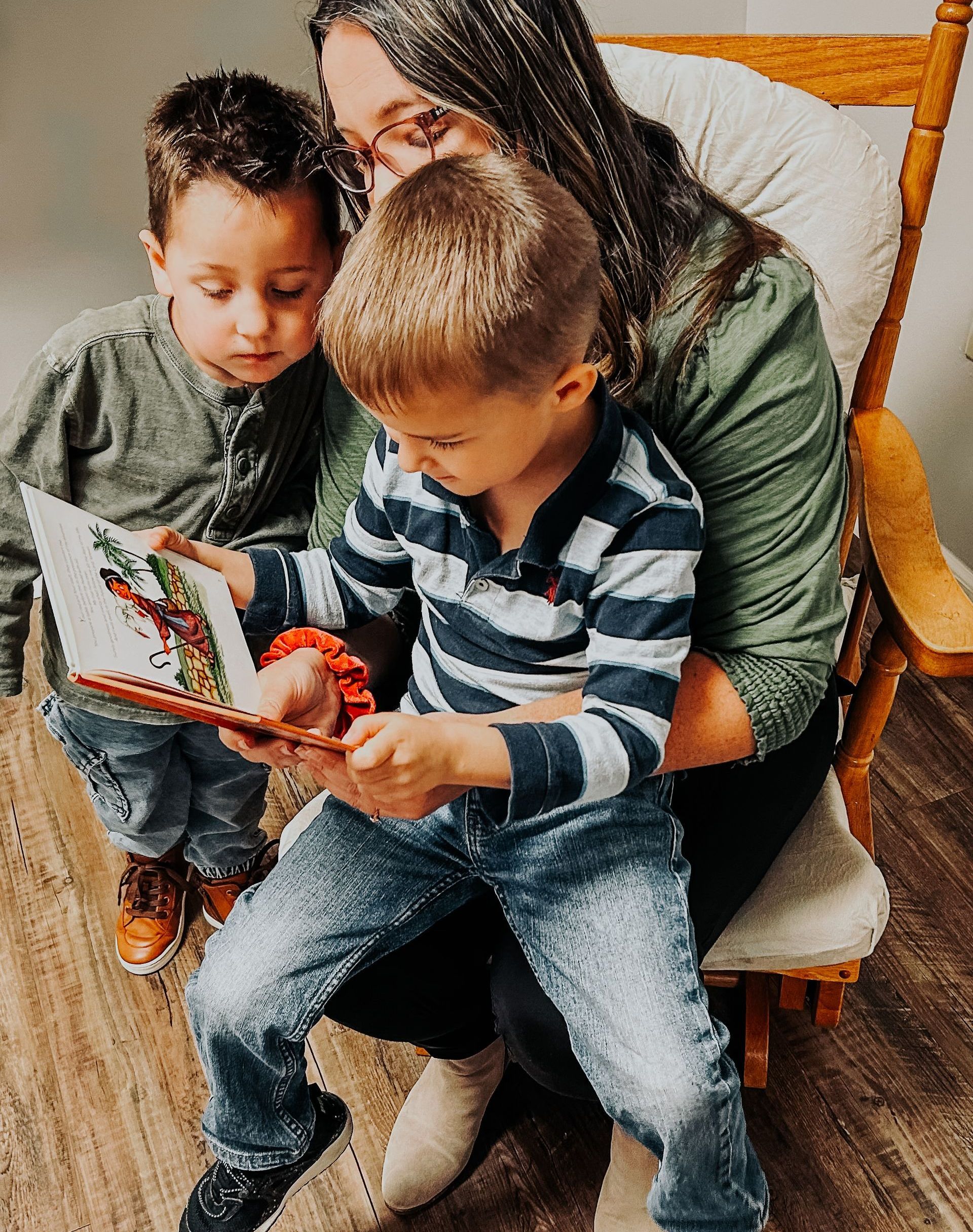 A person sits in a wooden rocking chair reading a book to two children.