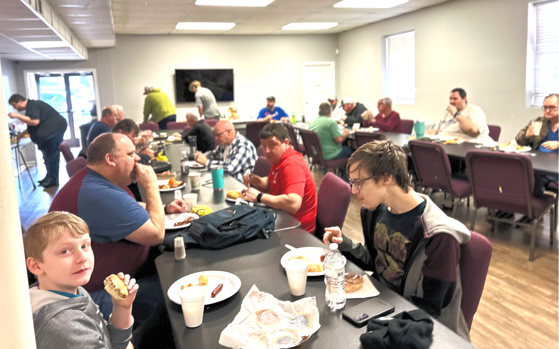 A group of people eating lunch together in a bright, cafeteria-style room with rows of tables and chairs.