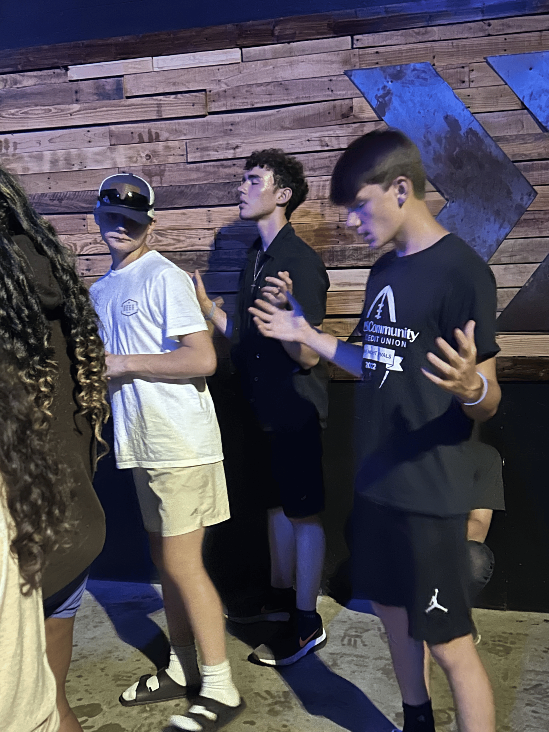 Three people stand in front of a wood-paneled wall, two with hands raised in a prayer-like gesture.
