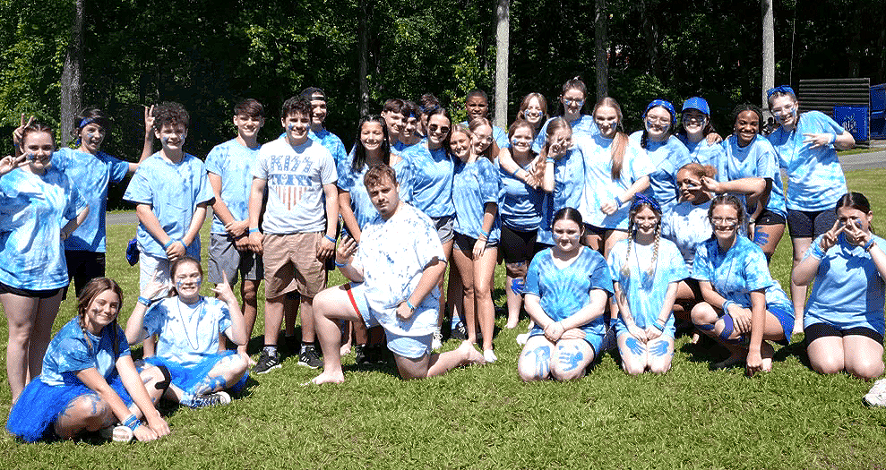 A large group of people in matching light blue t-shirts pose for a photo in a sunny, tree-lined outdoor setting.