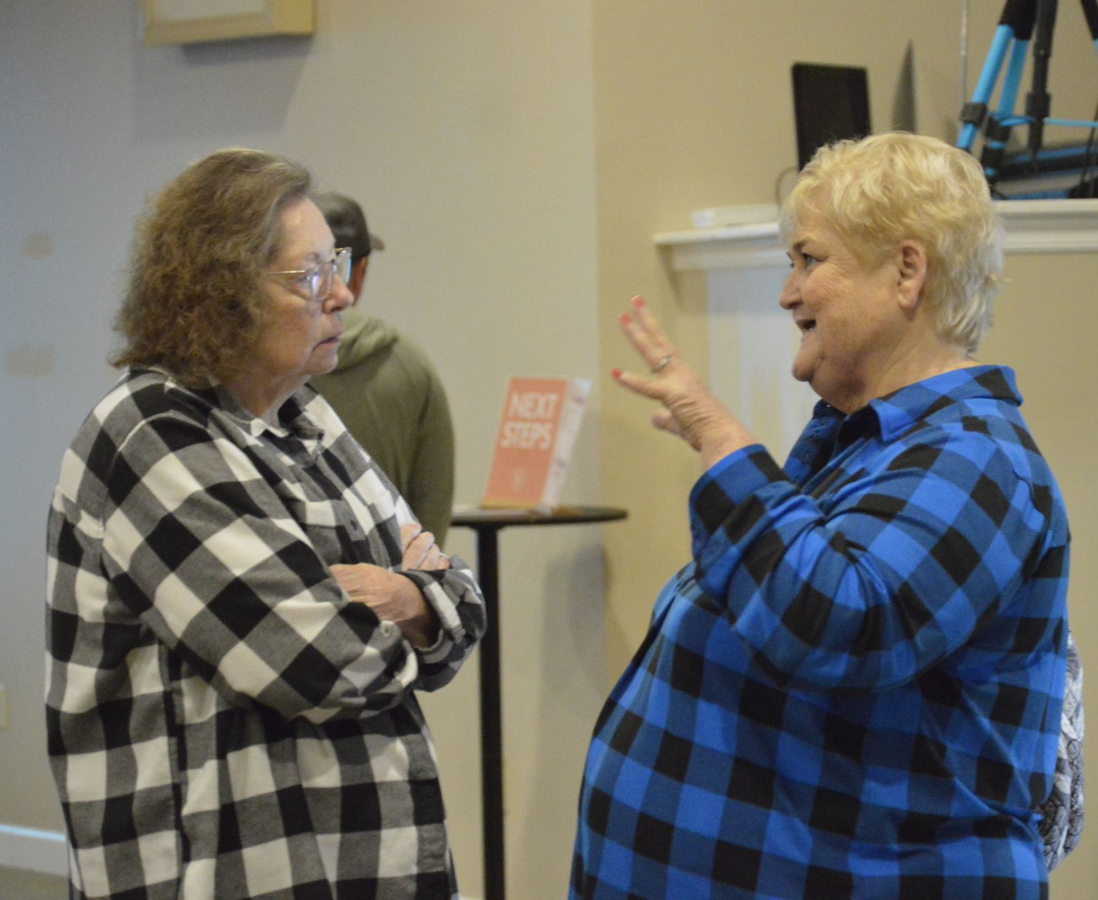 Two people in plaid shirts stand talking, one gesturing while the other listens with arms crossed, near a 'Next Steps' sign.