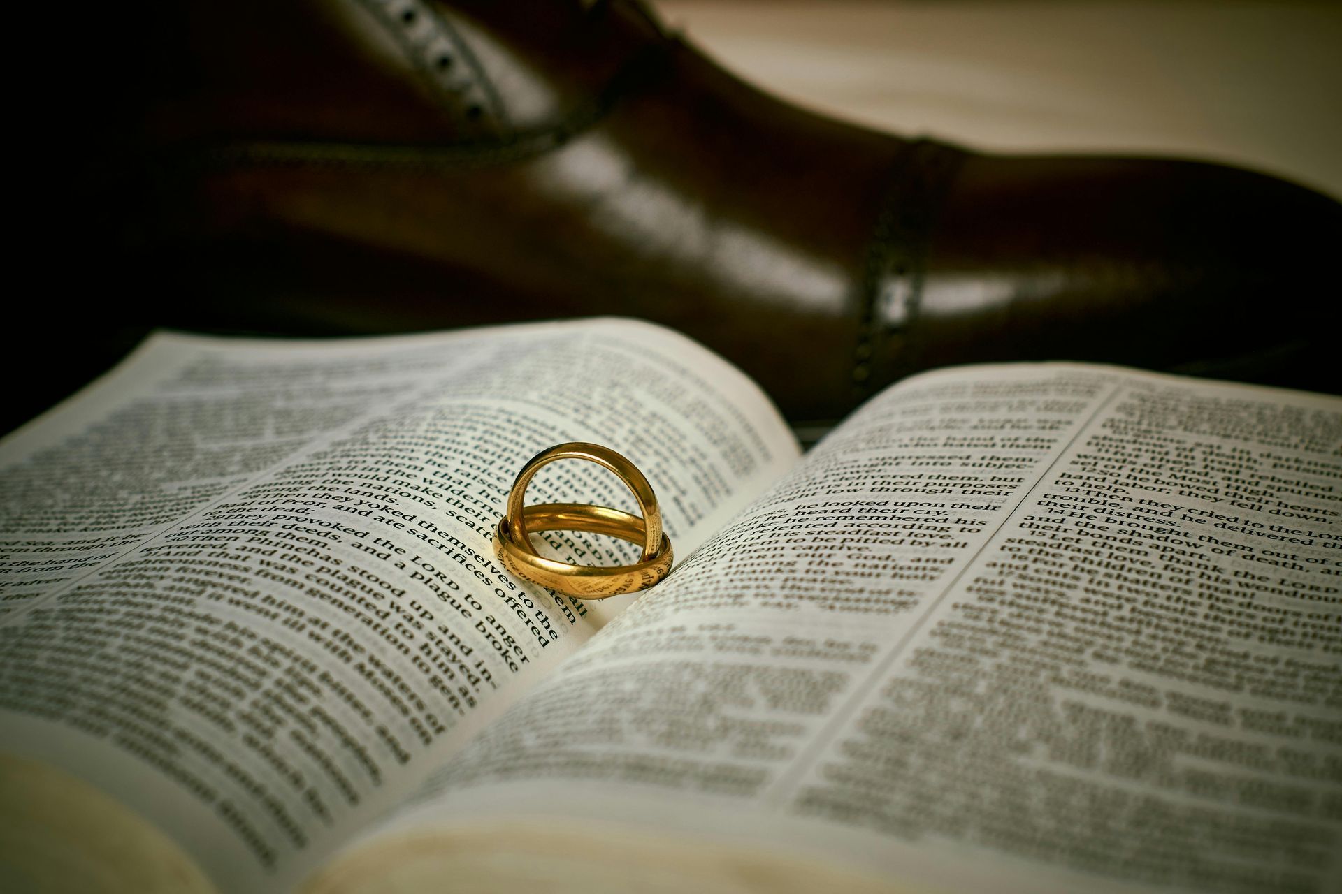 Two gold wedding bands resting on the pages of an open book with a formal leather shoe in the background.