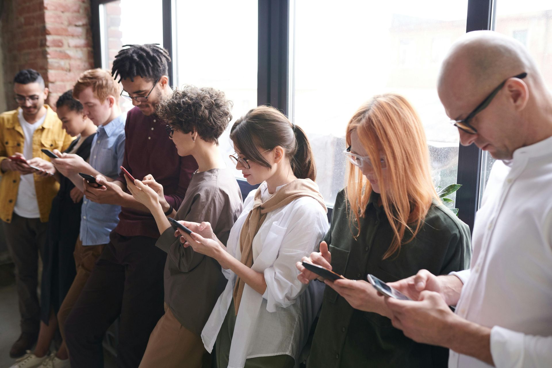 A group of people standing in a line indoors, all looking down at their smartphones.
