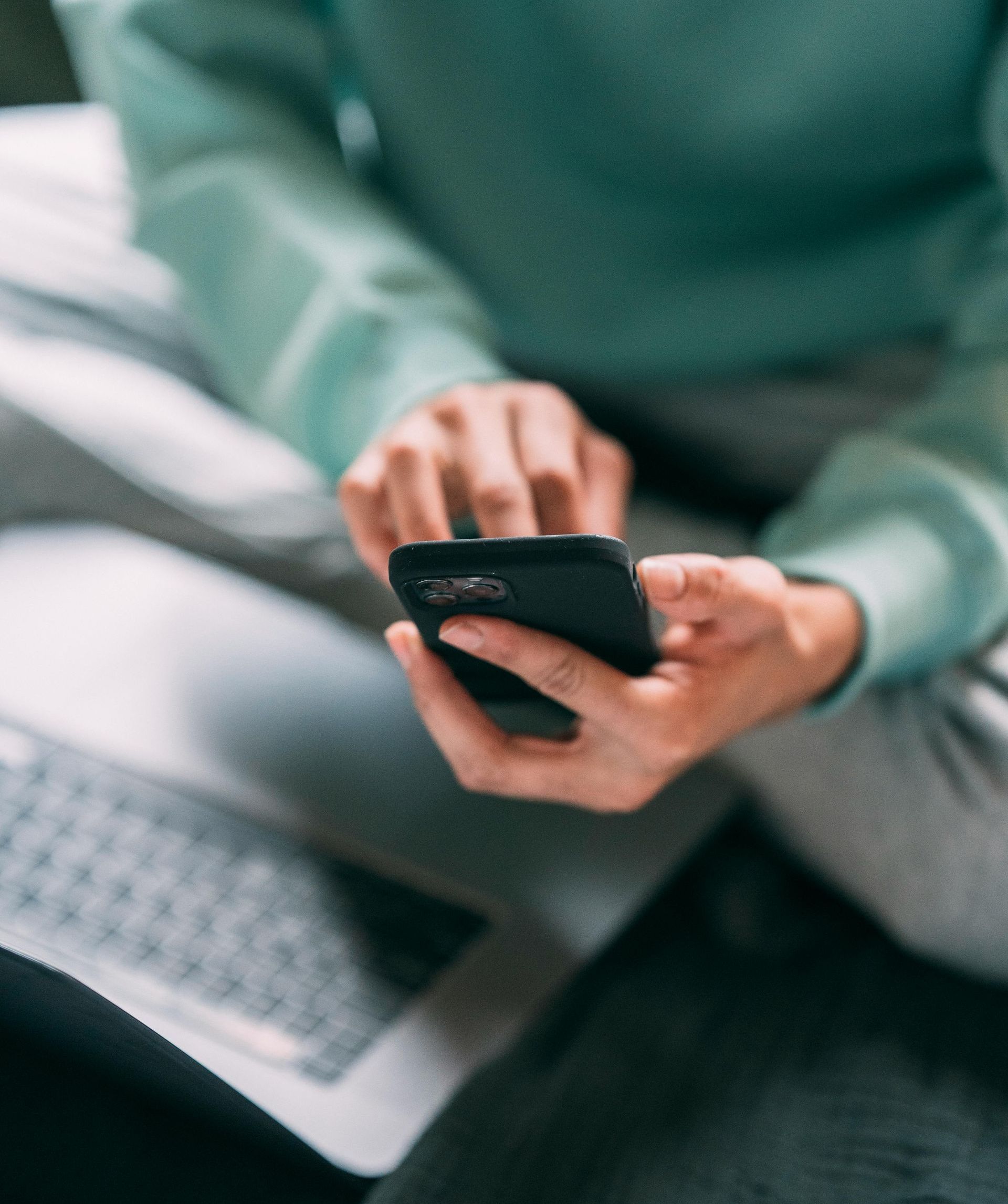 A person in a mint green sweater holds a black smartphone, with a laptop keyboard partially visible in the foreground.