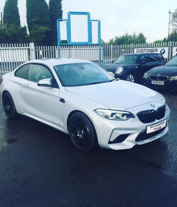 Silver BMW M2 coupe parked on wet asphalt, near a dealership, with other cars in the background.