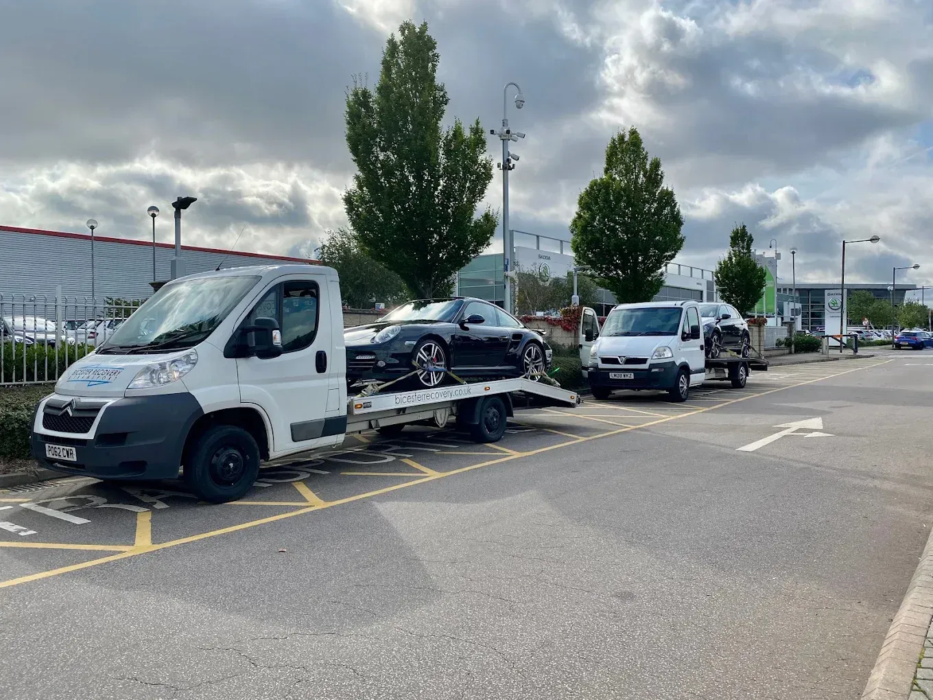 Two tow trucks parked on a road, one carrying a black car. Cloudy sky in the background.