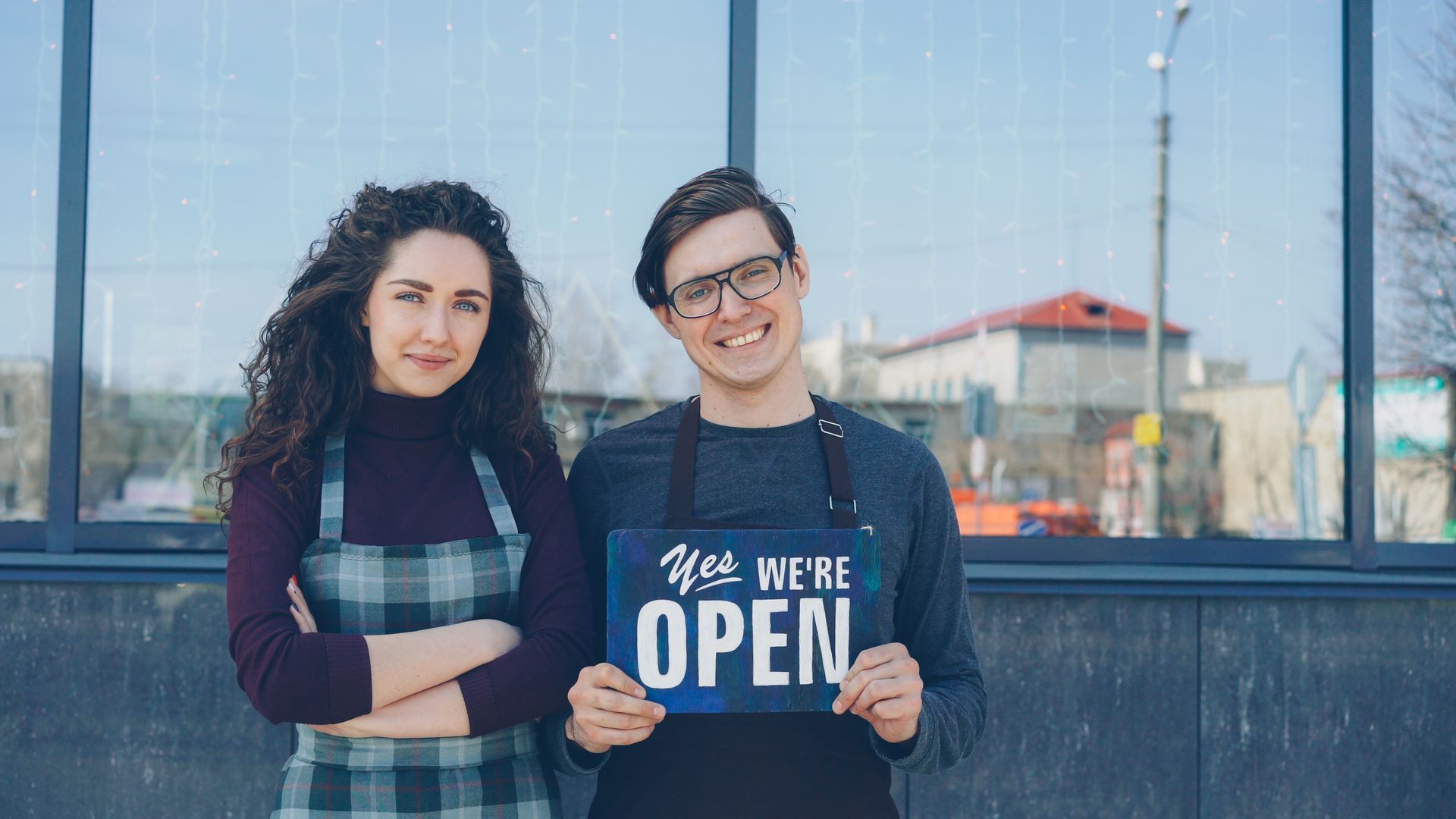Two small business owners smiling outside their storefront holding a sign that says we are open