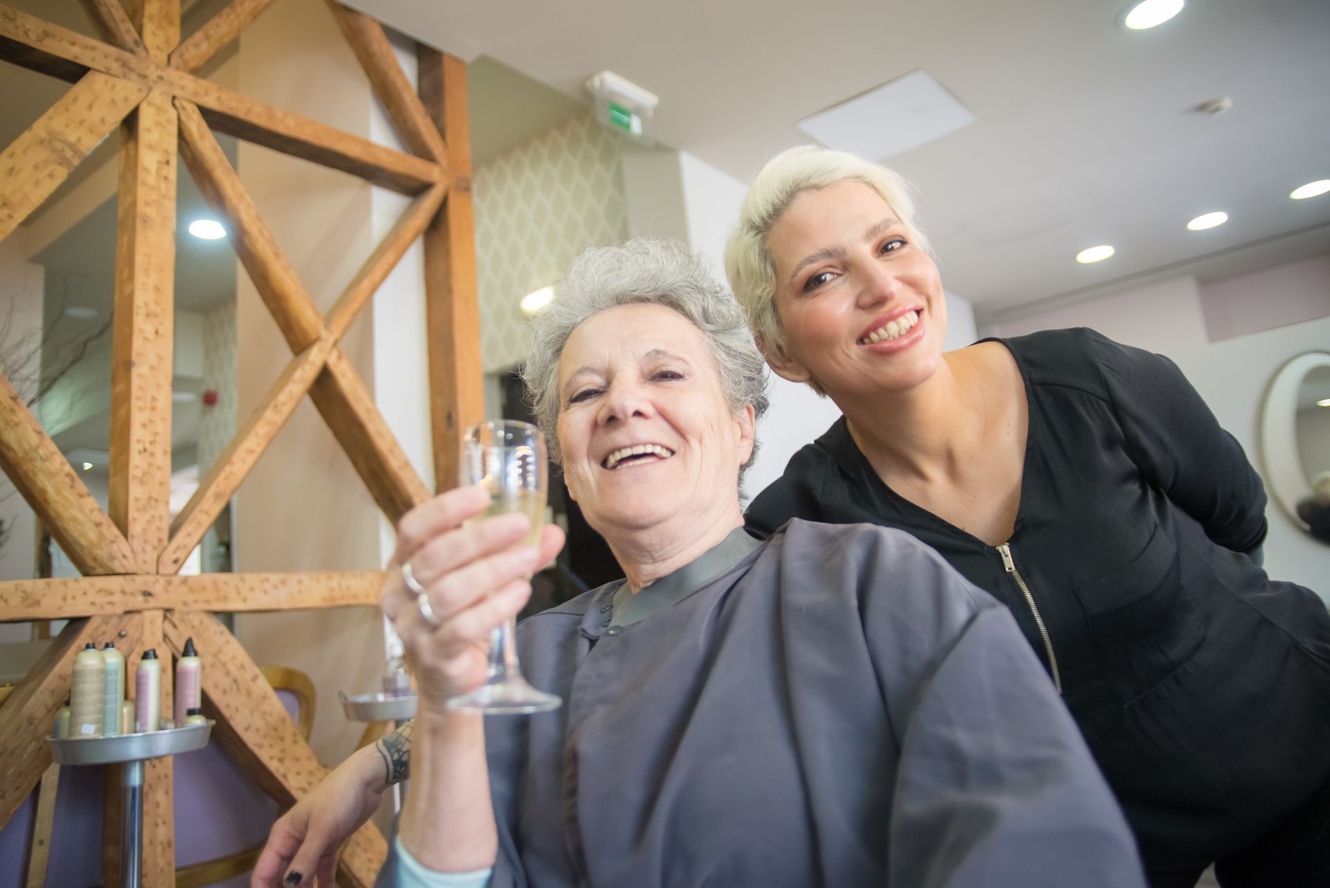 Smiling woman posing with an elderly resident in a bright care facility, holding a glass.