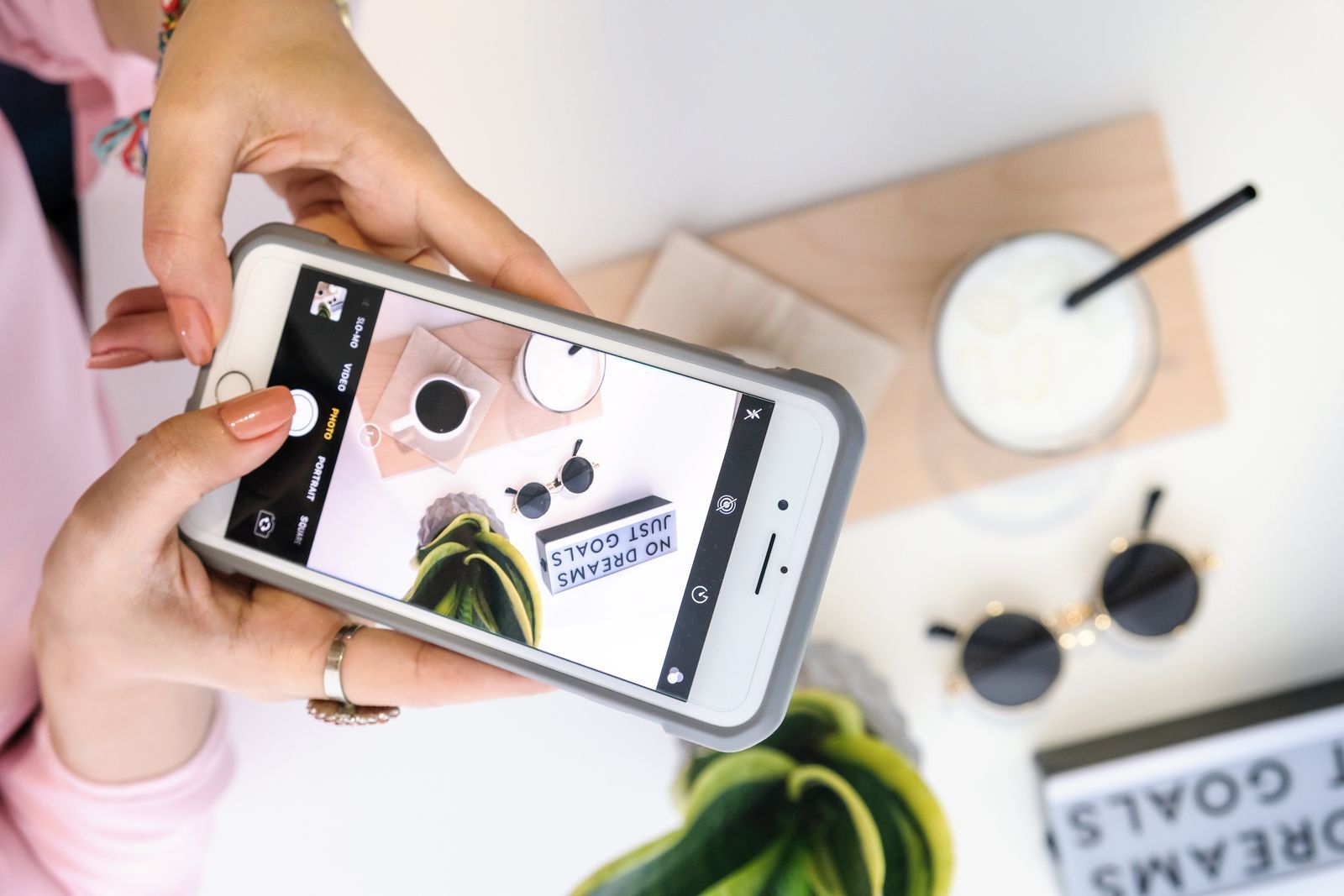 Hands holding a phone photographing coffee and sunglasses on a white desk