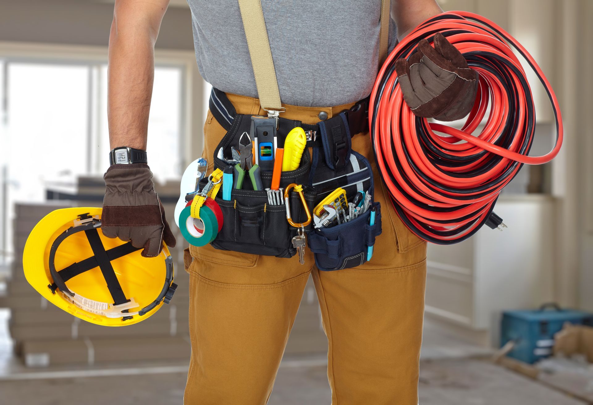 Electrician wearing tool belt and holding a coiled orange extension cord and yellow hard hat in an interior setting.