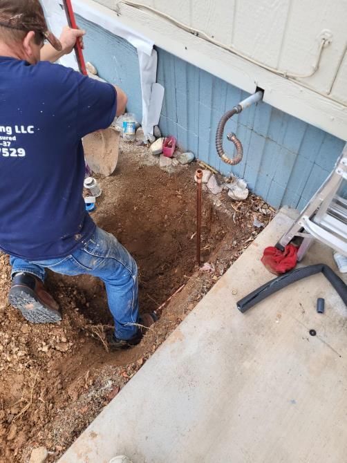 Man digging a hole next to a blue house foundation with a curved copper pipe visible.
