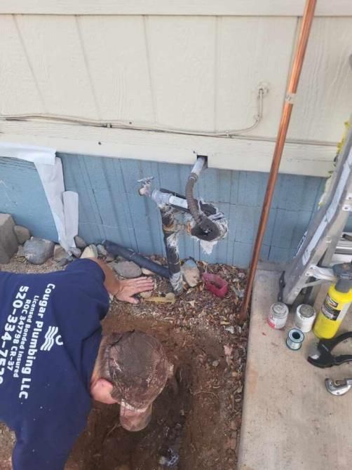 Plumber working on outdoor pipes; digging and connecting, with tools and materials visible. Light blue siding.
