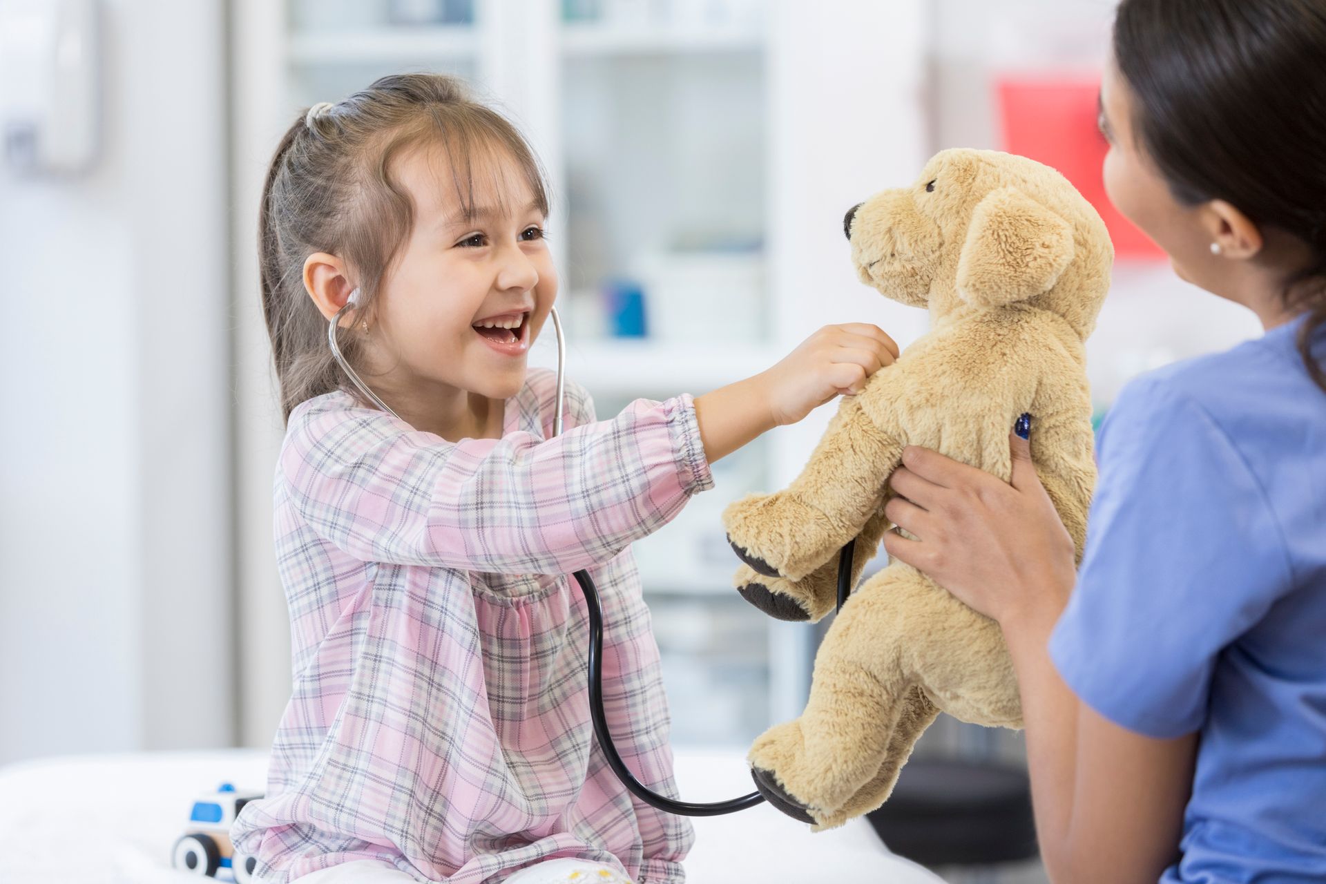 Child smiling at a teddy bear with a clinician in a bright medical exam room