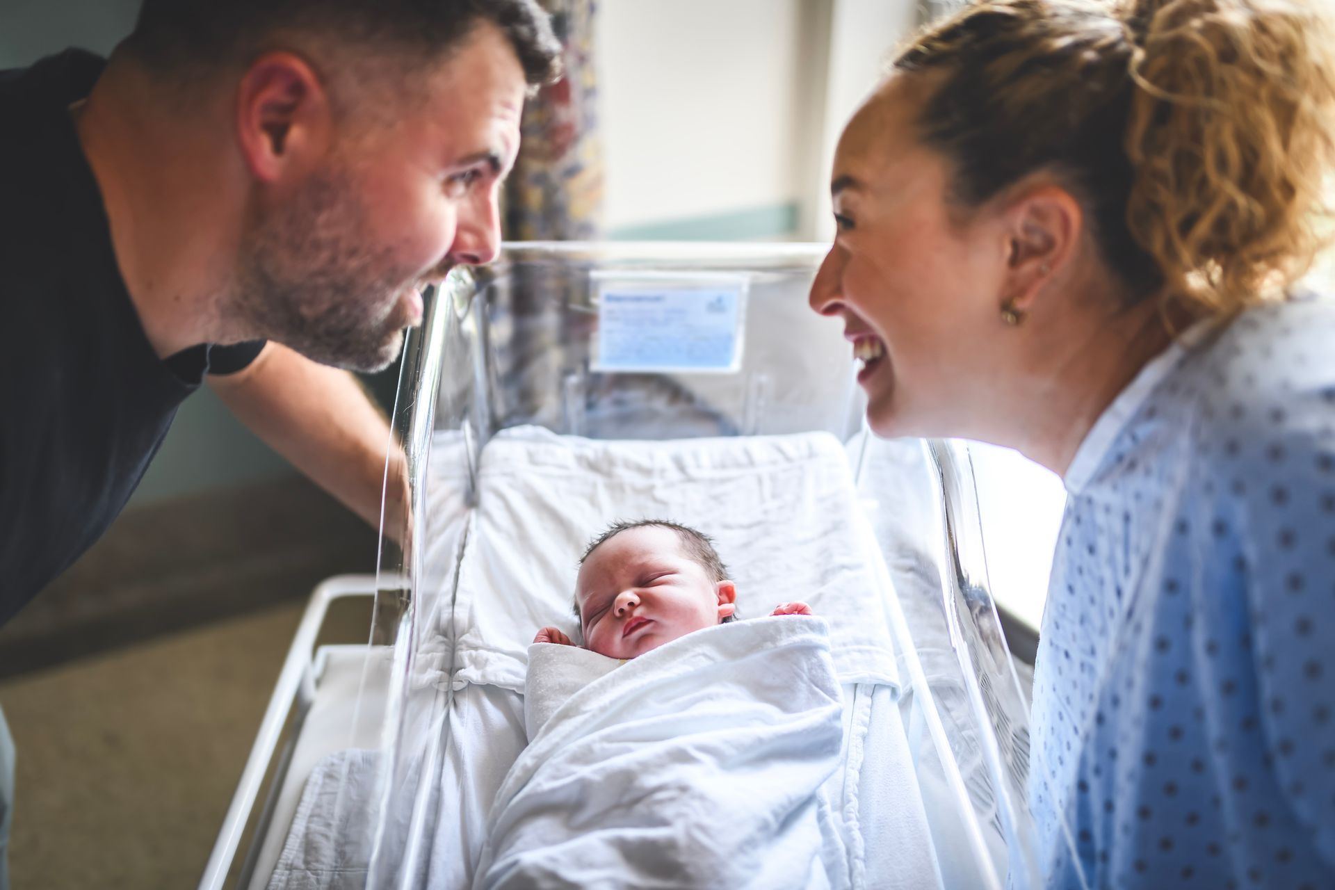 Parents look adoringly at newborn baby in hospital bassinet, smiling.