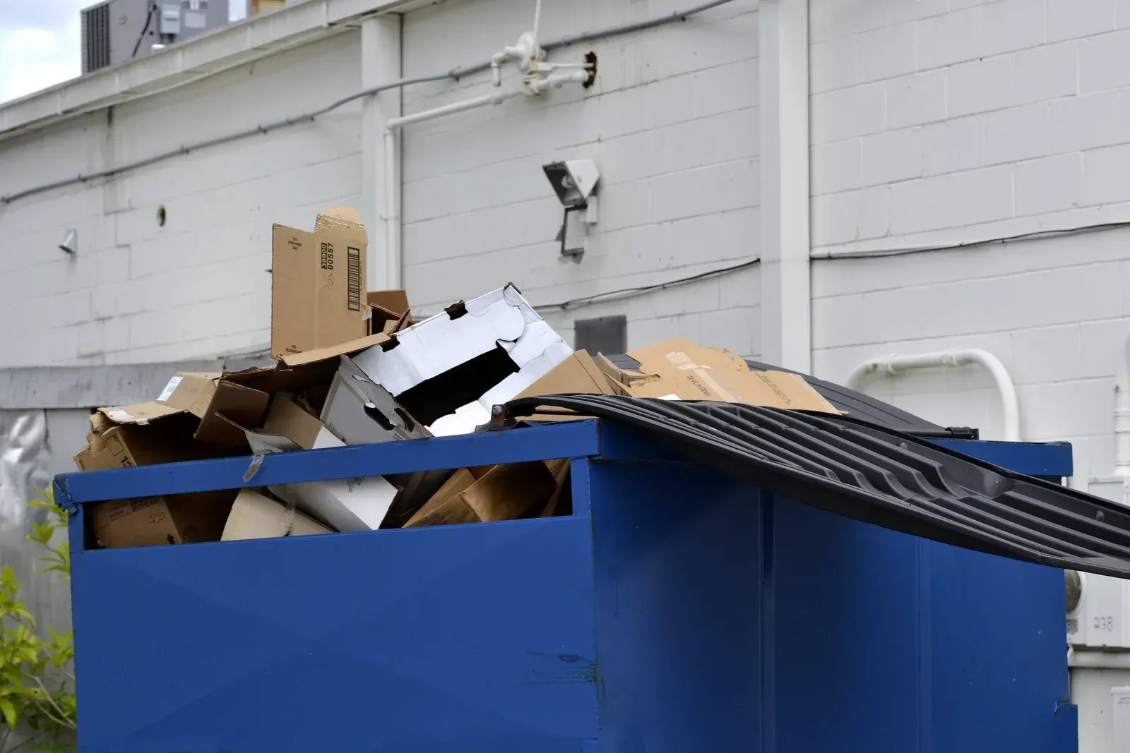 Blue dumpster overflowing with cardboard boxes near a white building.