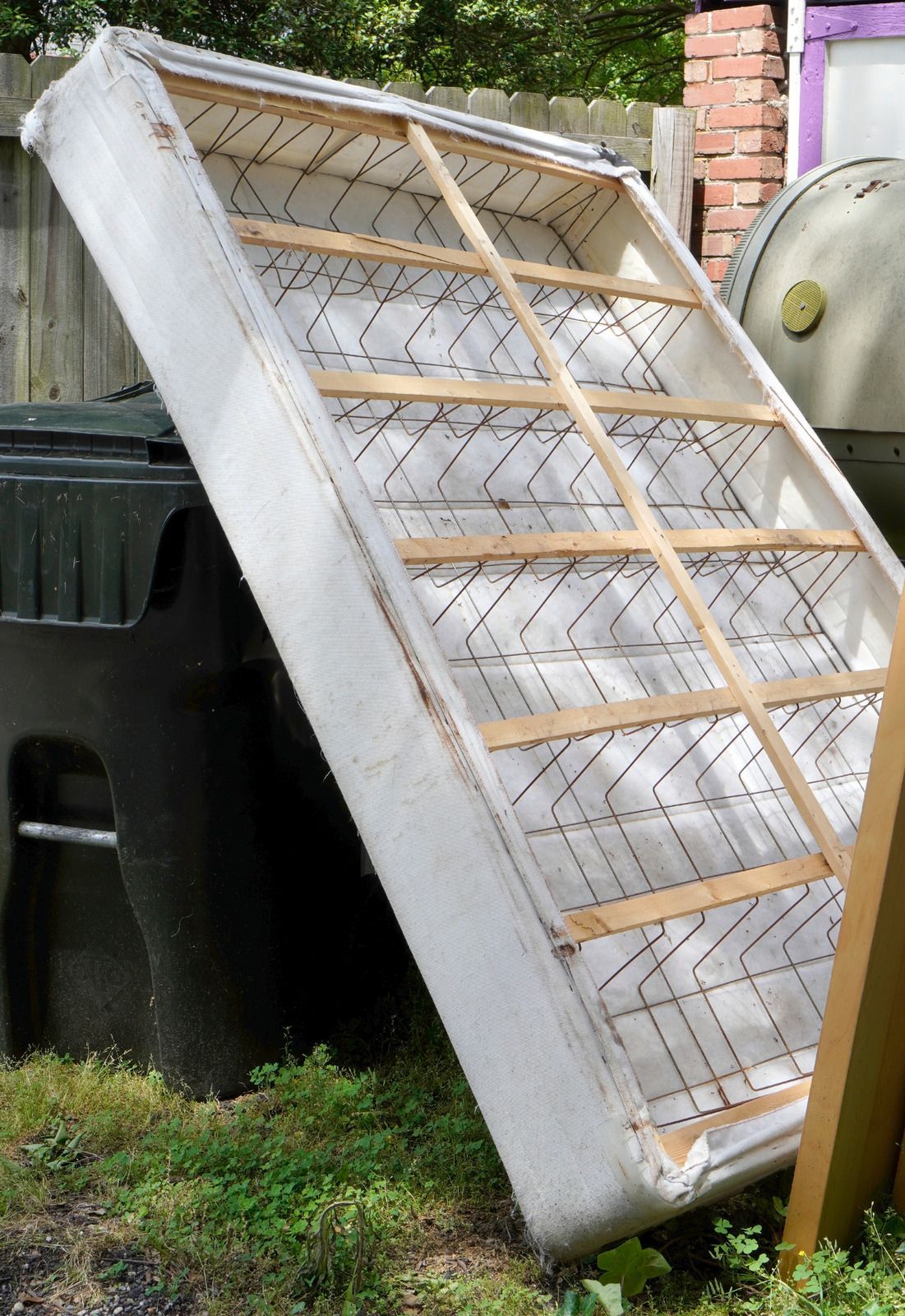 An old white mattress propped up against a trash can outdoors. Worn springs and wooden frame are visible.