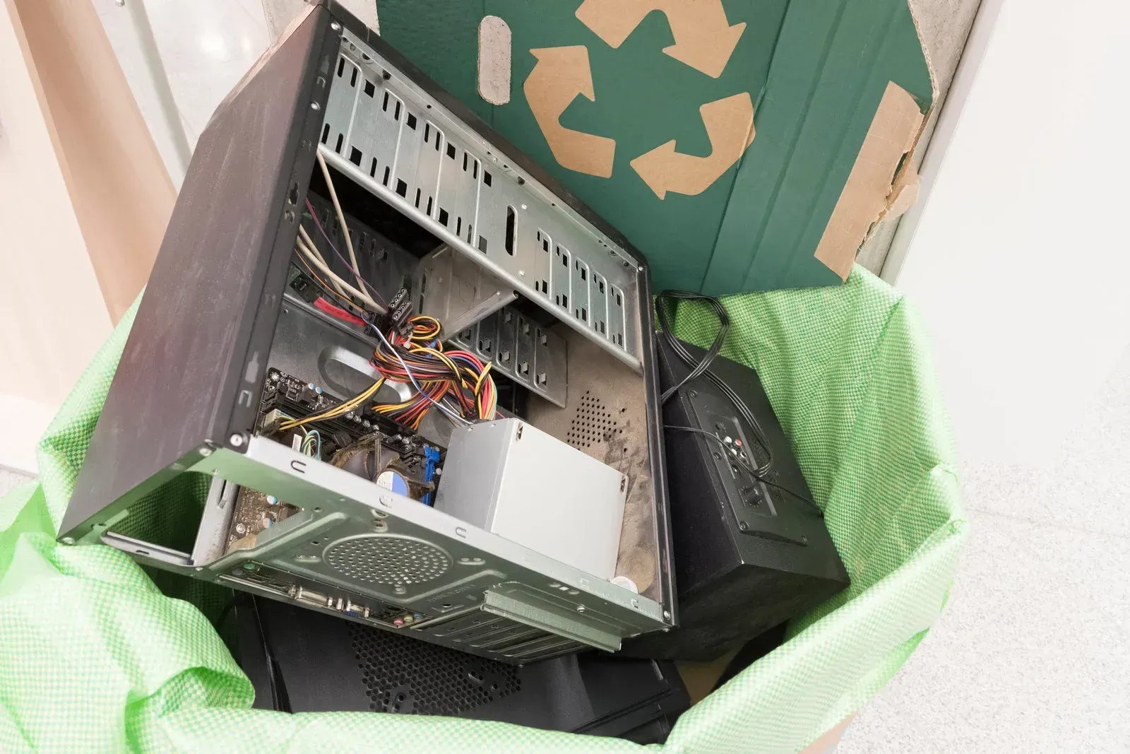 Person holding a green bag filled with discarded computer parts, including a desktop tower, with a recycle symbol visible.