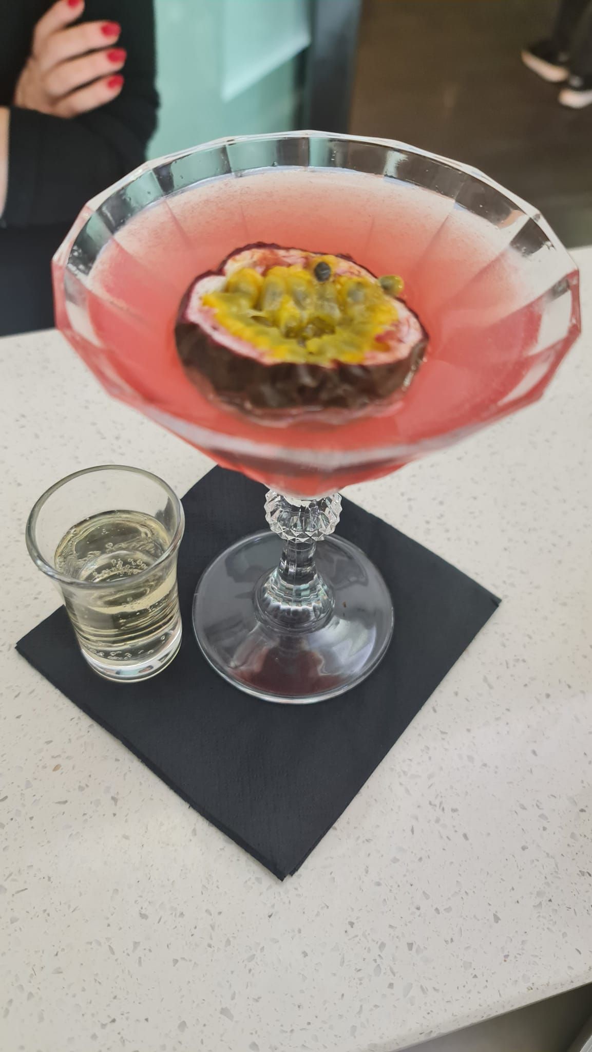 Cocktail rosa in un bicchiere da martini con guarnizione di frutto della passione, accanto a un bicchierino da shot su un tovagliolo.