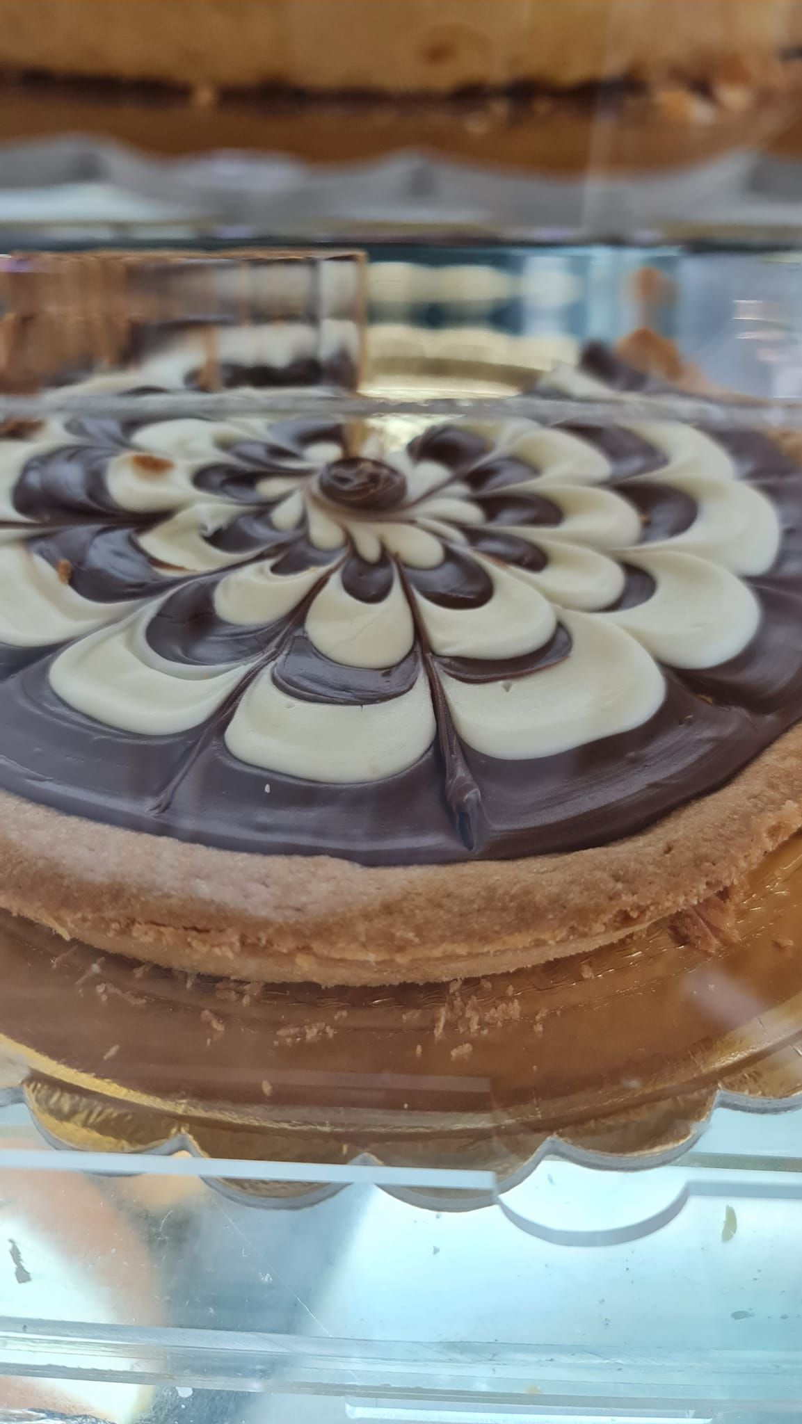Dessert rotondo con decorazione a spirale di cioccolato e fiori bianchi, servito su un piatto di vetro.