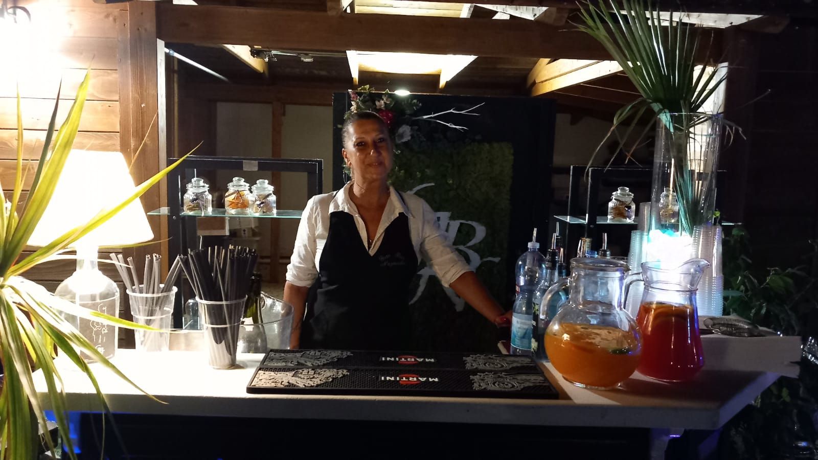 Un barista dietro al bancone, con bottiglie e bicchieri in un locale illuminato da una luce calda.