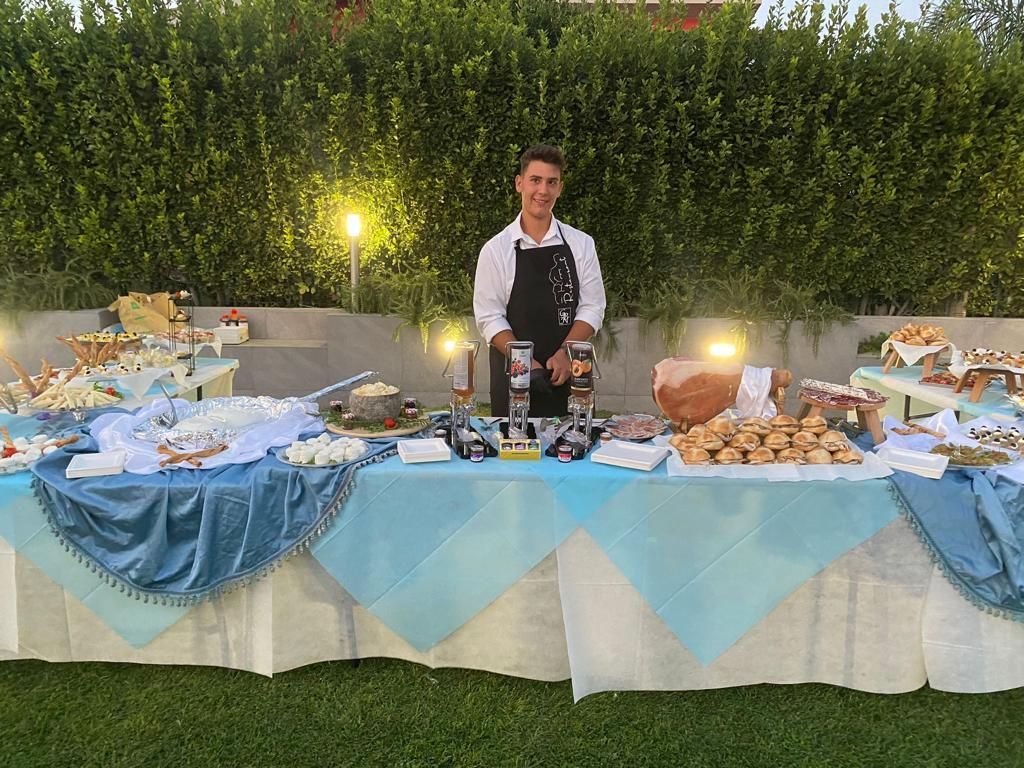 Persona in piedi dietro un tavolo da buffet con tovaglie blu durante un evento serale all'aperto.