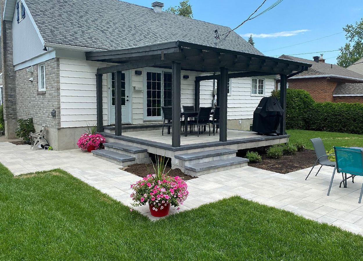 Patio arrière avec pergola, marches et pots de fleurs, relié à une maison blanche avec pavés gris et herbe verte.