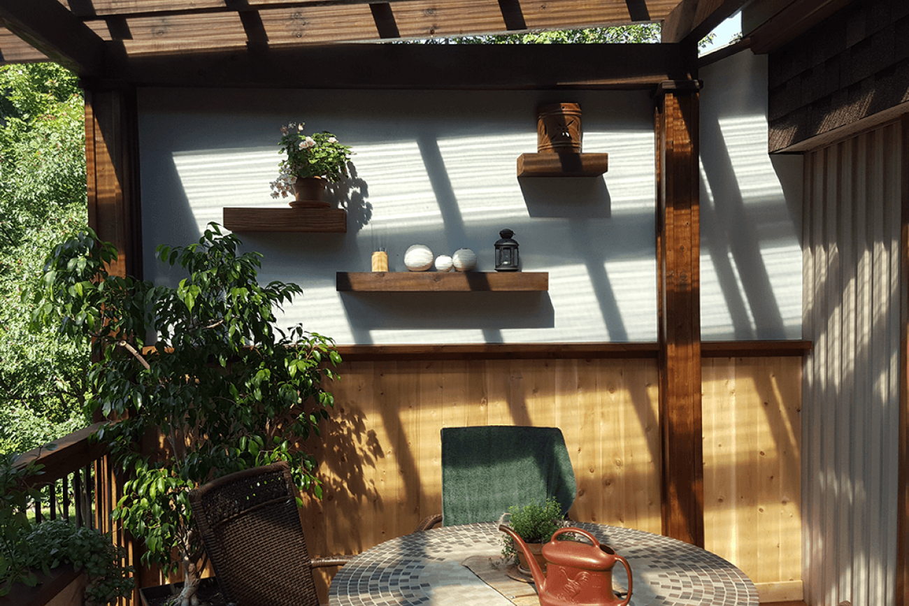 Terrasse avec étagères pour plantes et objets, pergola en bois marron, table avec housse à motifs, chaise verte.