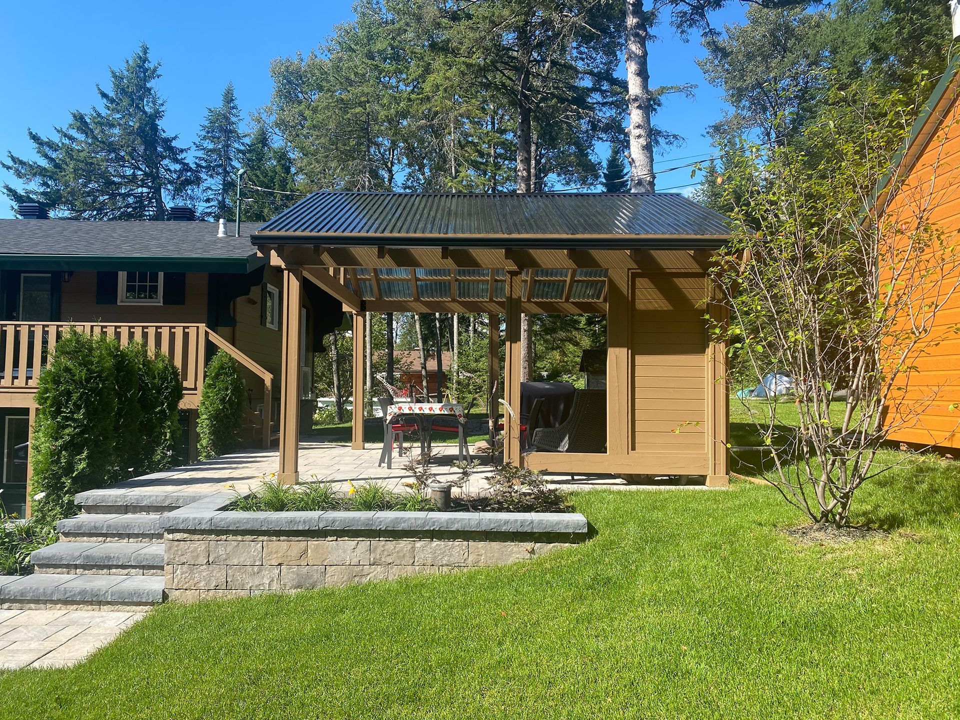 Abri barbecue en bois avec un toit transparent, sur une pelouse herbeuse avec un bâtiment en arrière-plan.