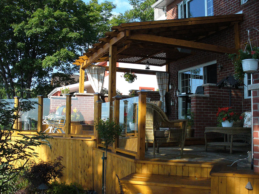 Terrasse en bois avec pergola, balustrade en verre et maison en briques. Coin salon extérieur avec plantes.