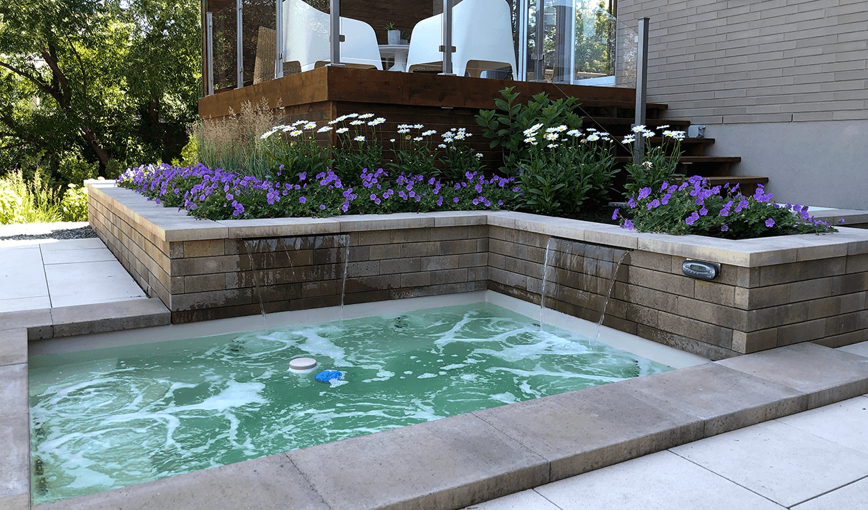 Bain à remous avec eau bouillonnante entouré de pierre, un jardin surélevé avec des fleurs et une terrasse.