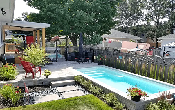 Jardin avec piscine rectangulaire, terrasse et coin détente. On aperçoit des chaises rouges, de la verdure et une pergola.