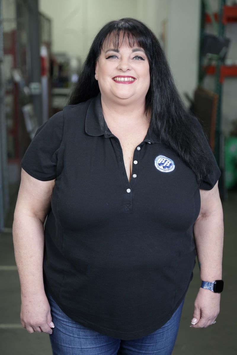 Woman in a black polo shirt standing and smiling in a workshop doorway.