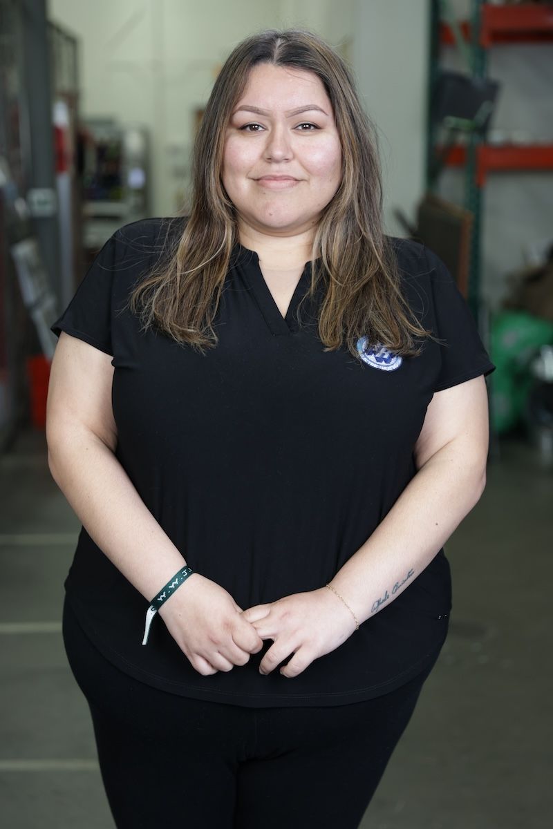 Woman standing in a workshop, wearing a black polo and pants, with a slight smile.