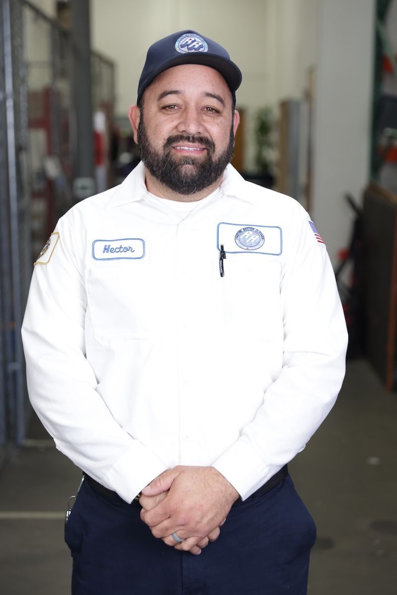 Man in white uniform and navy cap standing indoors with hands clasped, smiling at the camera