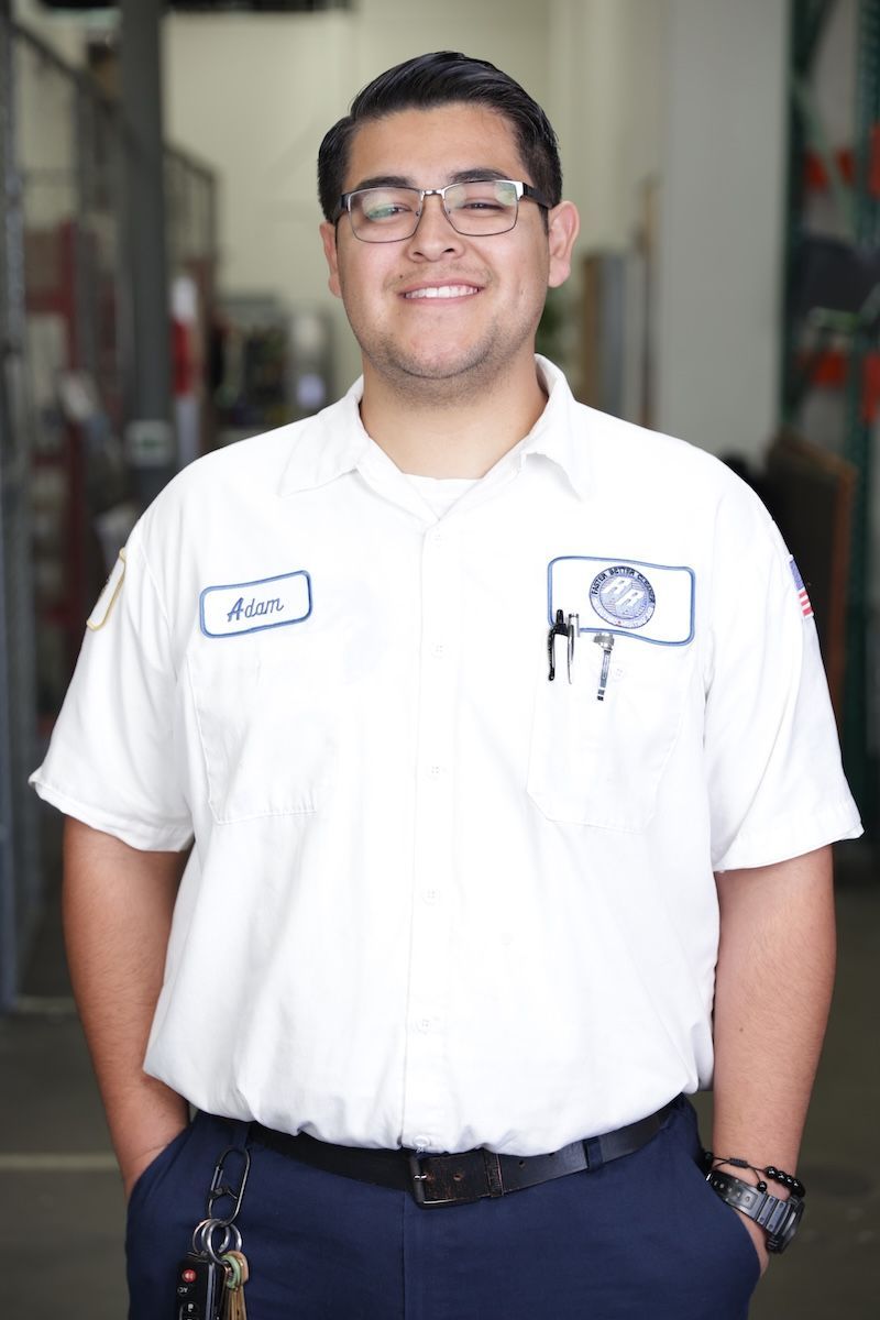 Man in a white work uniform standing in a workshop doorway, smiling and holding a phone in a shirt pocket.