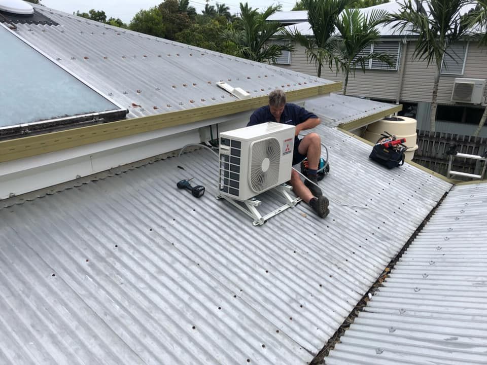 A Man Is Sitting On The Roof Of A House Fixing An Air Conditioner — Beards Refrigeration & Air Conditioning in Mackay, QLD