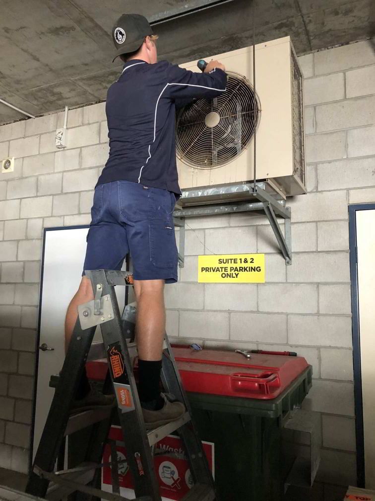 A Man Is Standing On A Ladder Fixing An Air Conditioner — Beards Refrigeration & Air Conditioning in Bucasia, QLD